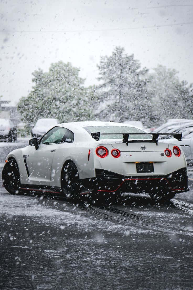 白色Nissan R35 GT-R雪地行駛圖 壁紙 — 一輛白色日產GT-R跑車，紅色尾燈，在濕滑停車場中穿越大雪行駛。