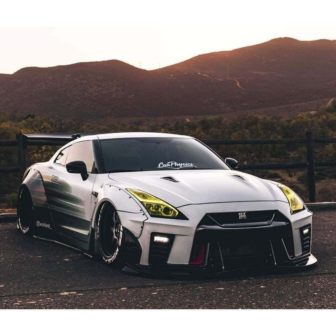 白色Nissan R35 GT-R山景圖 壁紙 — 一架白色改裝日產GT-R跑車，配備寬體套件和黃色頭燈，停泊在瀝青路上，背景是日落時分的起伏山丘。