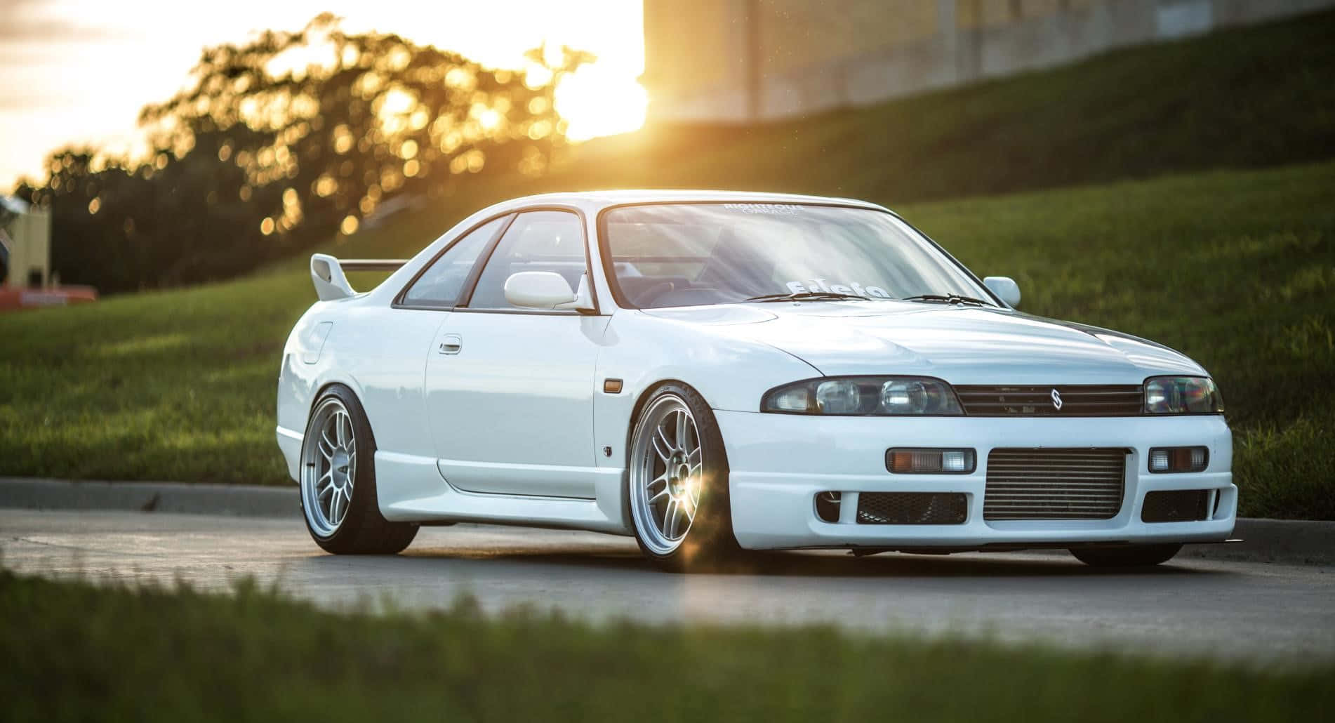 白色低調R33 GTR 壁紙 — 一輛純白色的日產Skyline R32跑車在夕陽金光下閃耀，行駛在兩旁長滿青草的道路上。