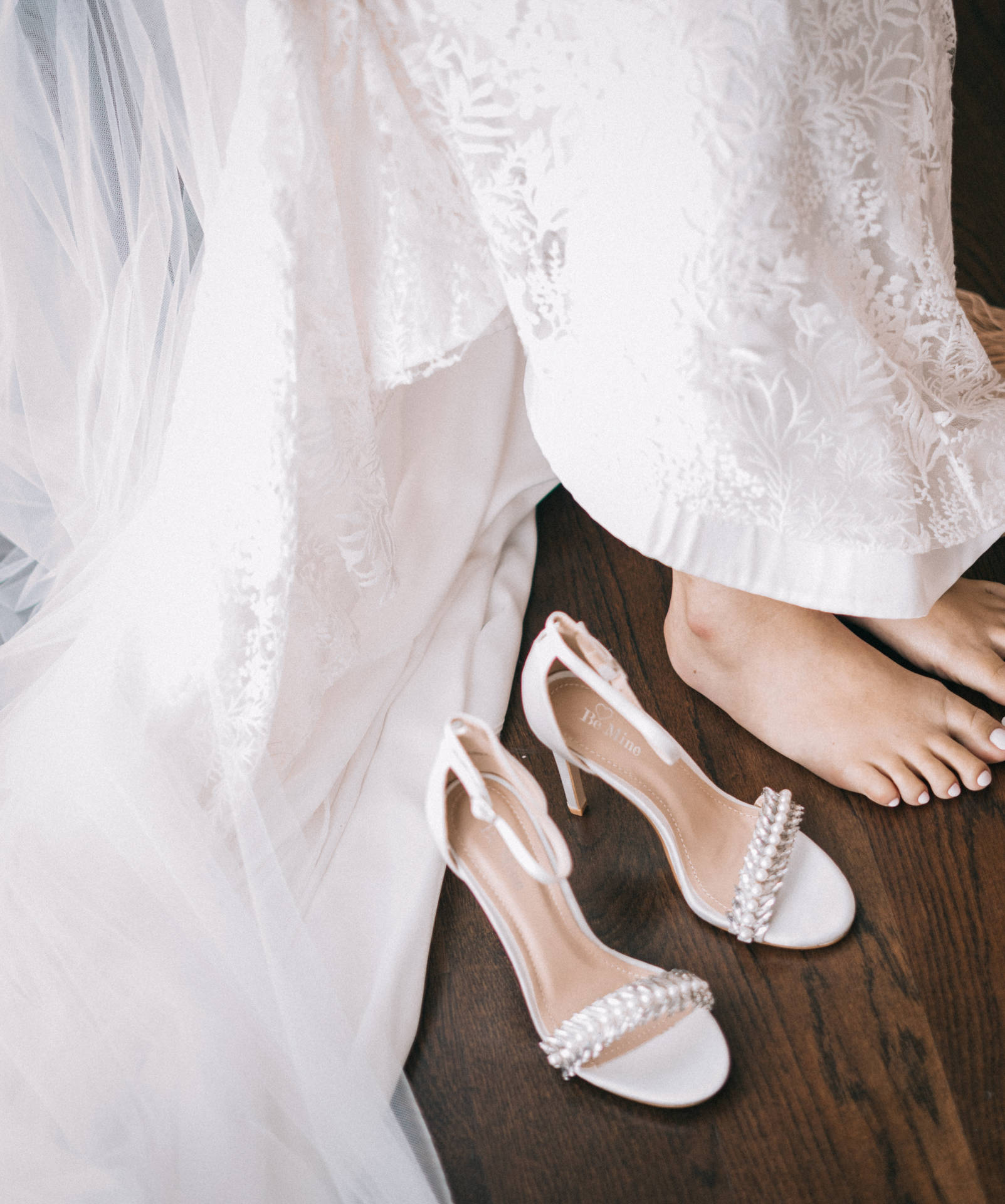 木地板上蕾絲婚紗旁邊的新娘赤腳和白色珠飾高跟鞋。