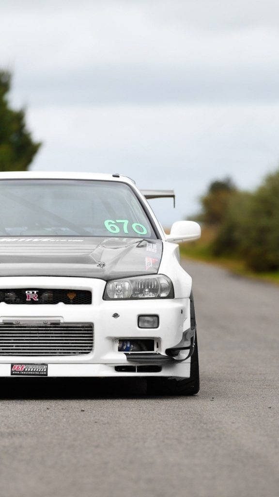 白色尼桑Skyline汽車 壁紙 — 一架白色日產Skyline R34賽車，擋風玻璃上有670號碼，停泊在鄉村瀝青路上。