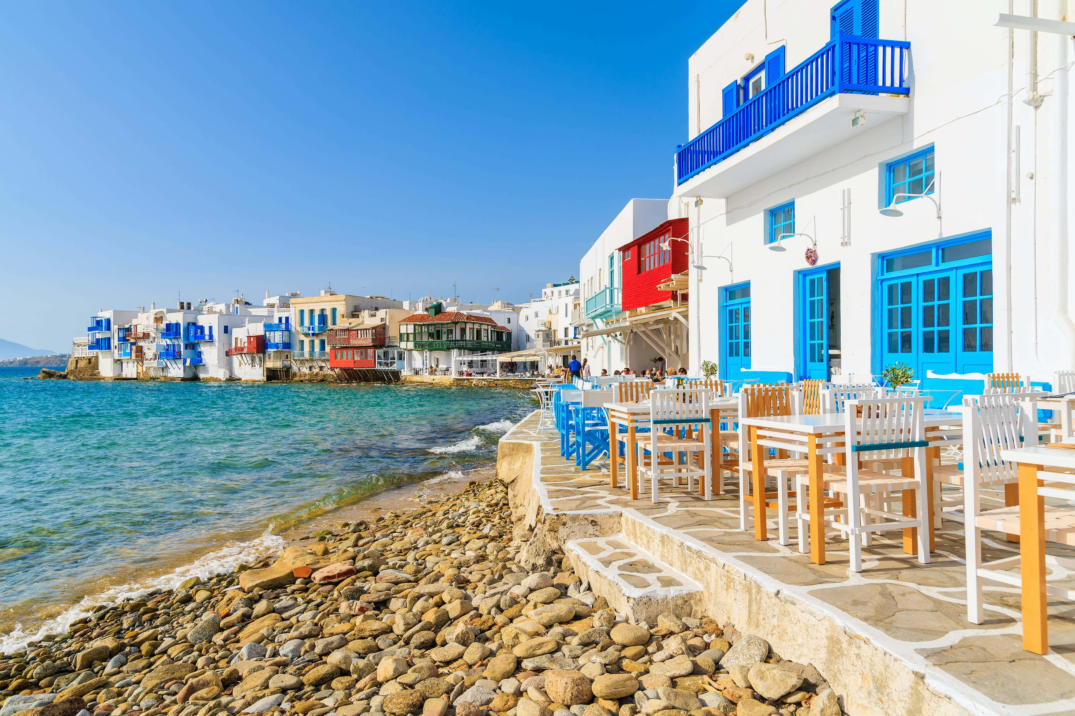 Greek Beach 桌布 壁紙 — 白色建築物配上大膽的藍色點綴，排列在一條陽光普照的海岸小徑旁，旁邊是碧綠的海水和卵石海灘。