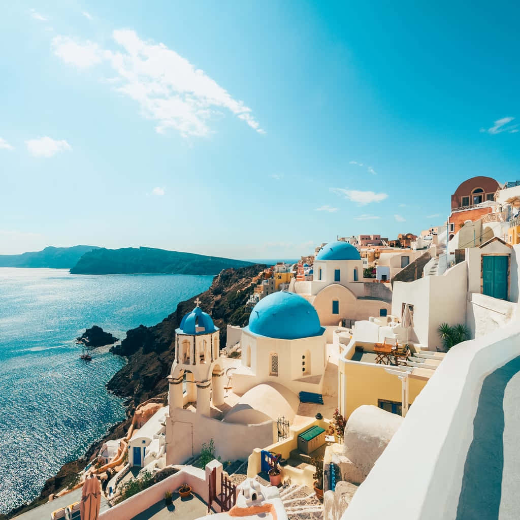 Greek Beach 桌布 壁紙 — 白色建築物配以鮮豔的藍色圓頂，戲劇性地棲息在懸崖上，俯瞰著清澈天空下深藍色的愛琴海。