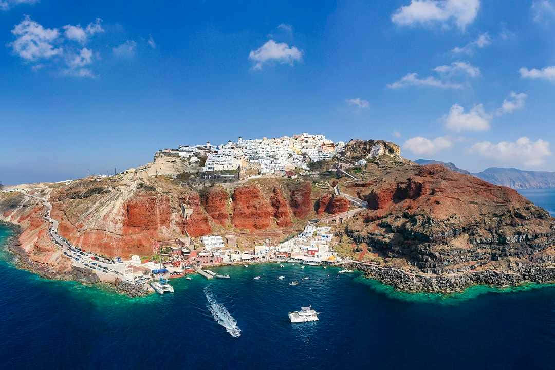 Greek Island 桌布 壁紙 — 白色懸崖村莊的鳥瞰圖，坐落在紅色岩石上，俯瞰著停泊船隻的碧綠海水。