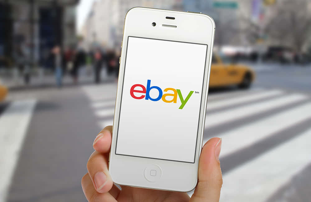 白色手機印有 eBay UK 標誌 壁紙 — 一隻手拿著白色iPhone，顯示色彩繽紛的eBay標誌，背景是模糊的城市街道和一輛黃色的士。