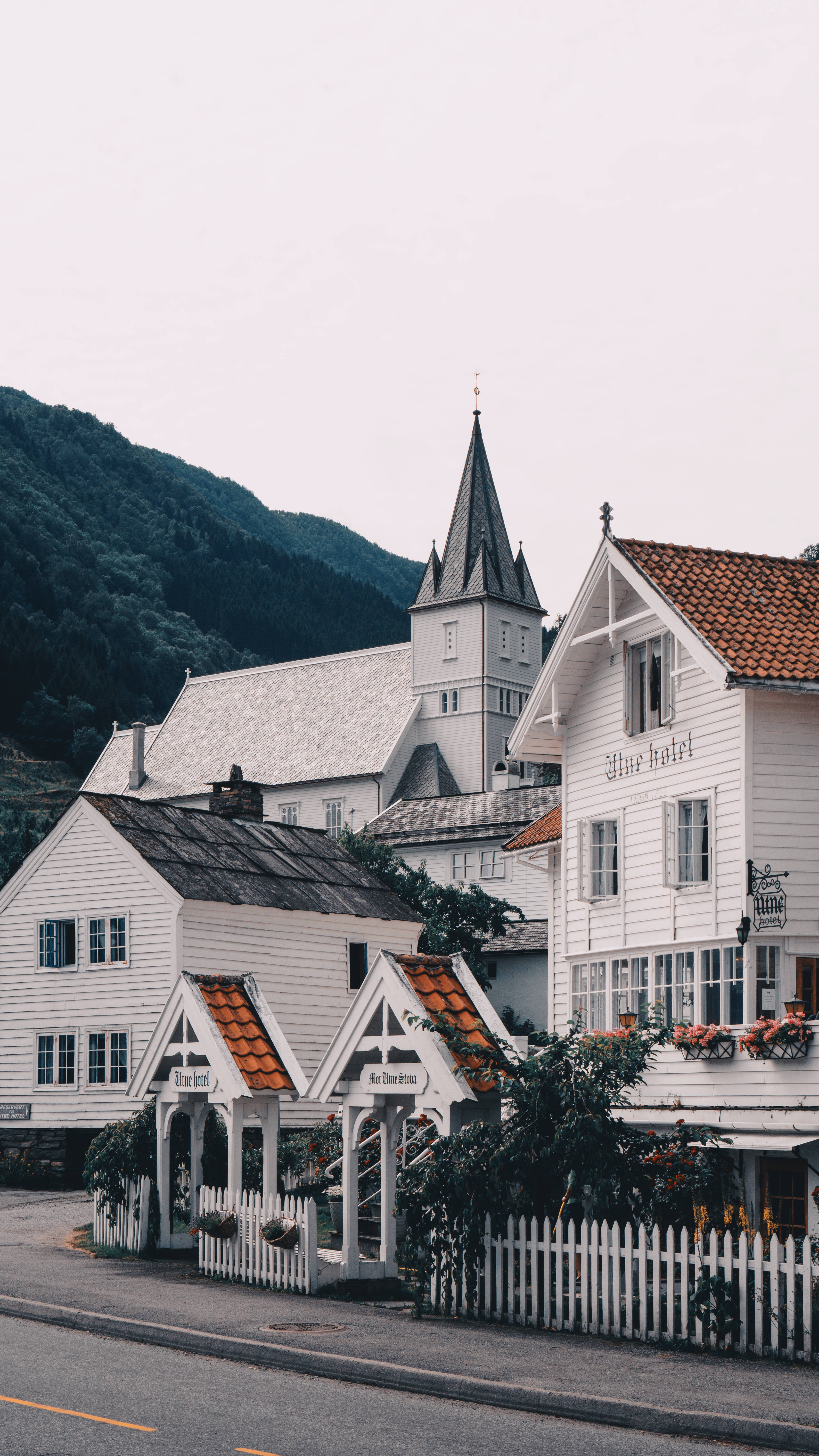 Norway 桌布 壁紙 — 白色木屋配紅瓦屋頂和教堂塔樓，依偎在森林覆蓋的山坡上，天空陰沉。