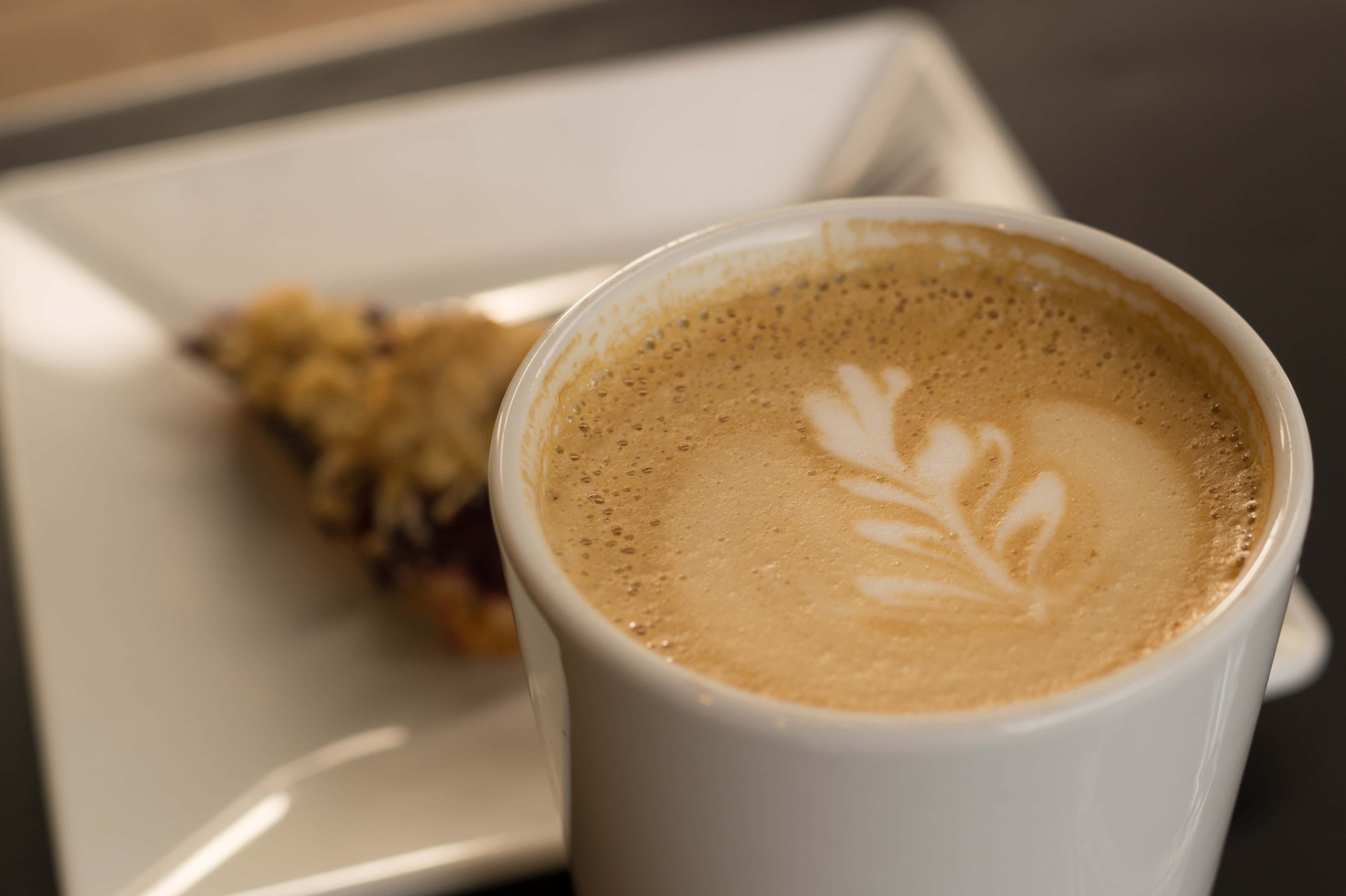 Coffee Cup 桌布 壁紙 — 白色杯子盛載咖啡，帶有精緻樹葉拉花藝術，旁邊碟子上有糕點的近距離特寫。