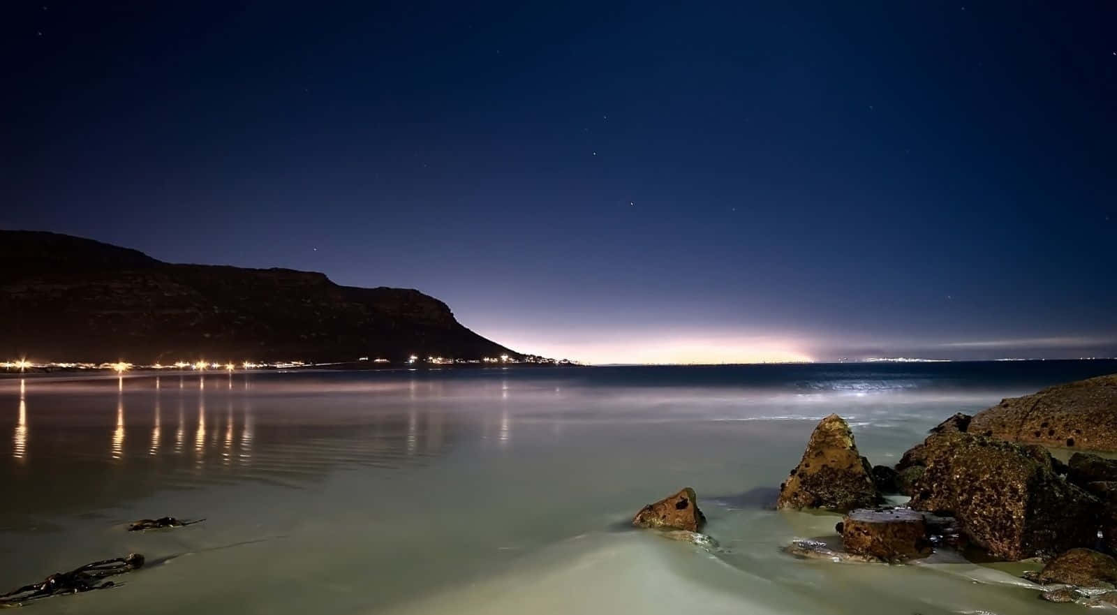 夜晚海灘，有星星、城市燈光同平靜嘅水面喺沙灘上反射。