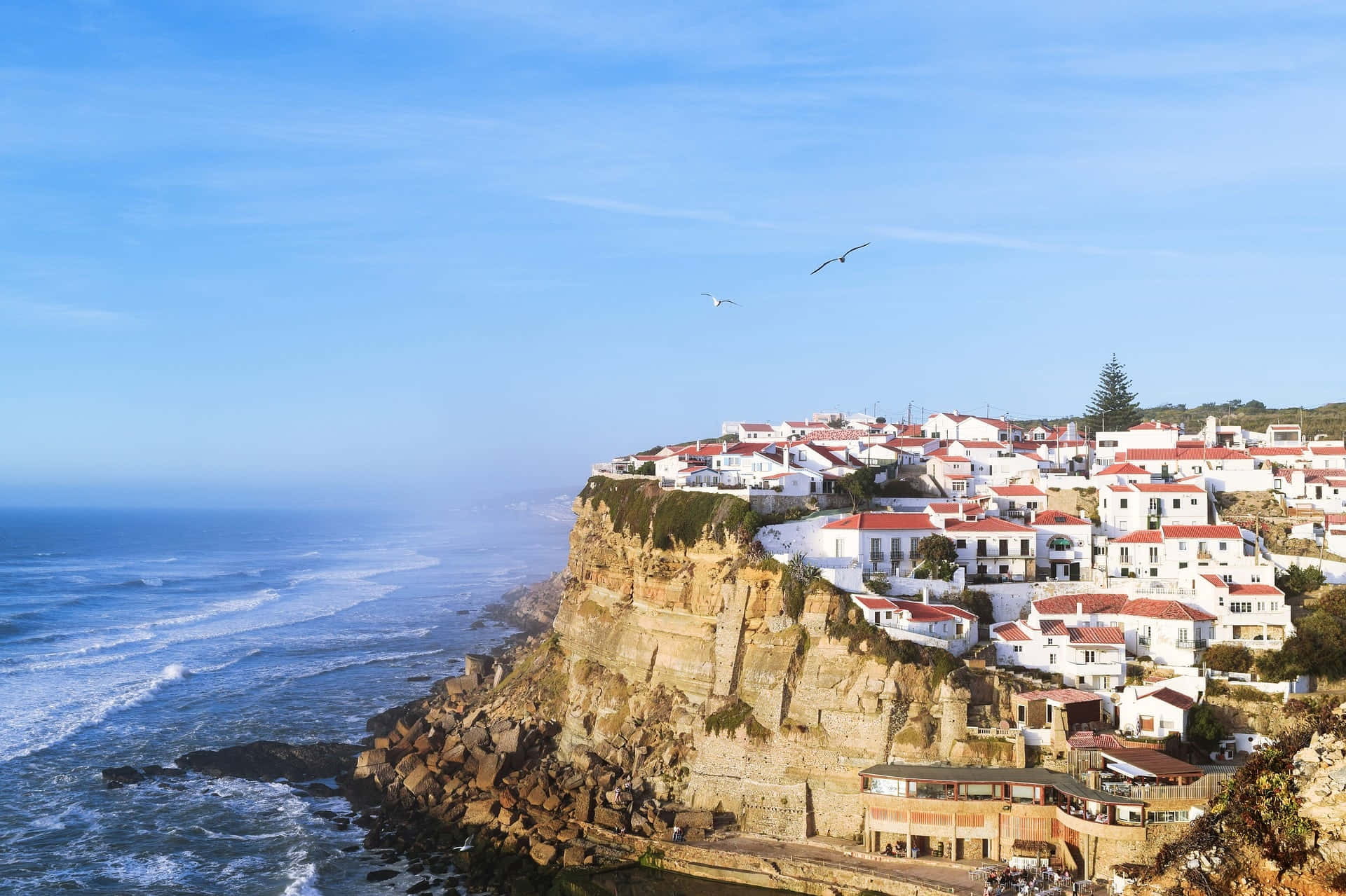 Portuguese Beach 桌布 壁紙 — 白色牆壁的村莊，紅色屋頂依附在崎嶇的懸崖上，俯瞰著清澈藍天下碧綠色的海洋。