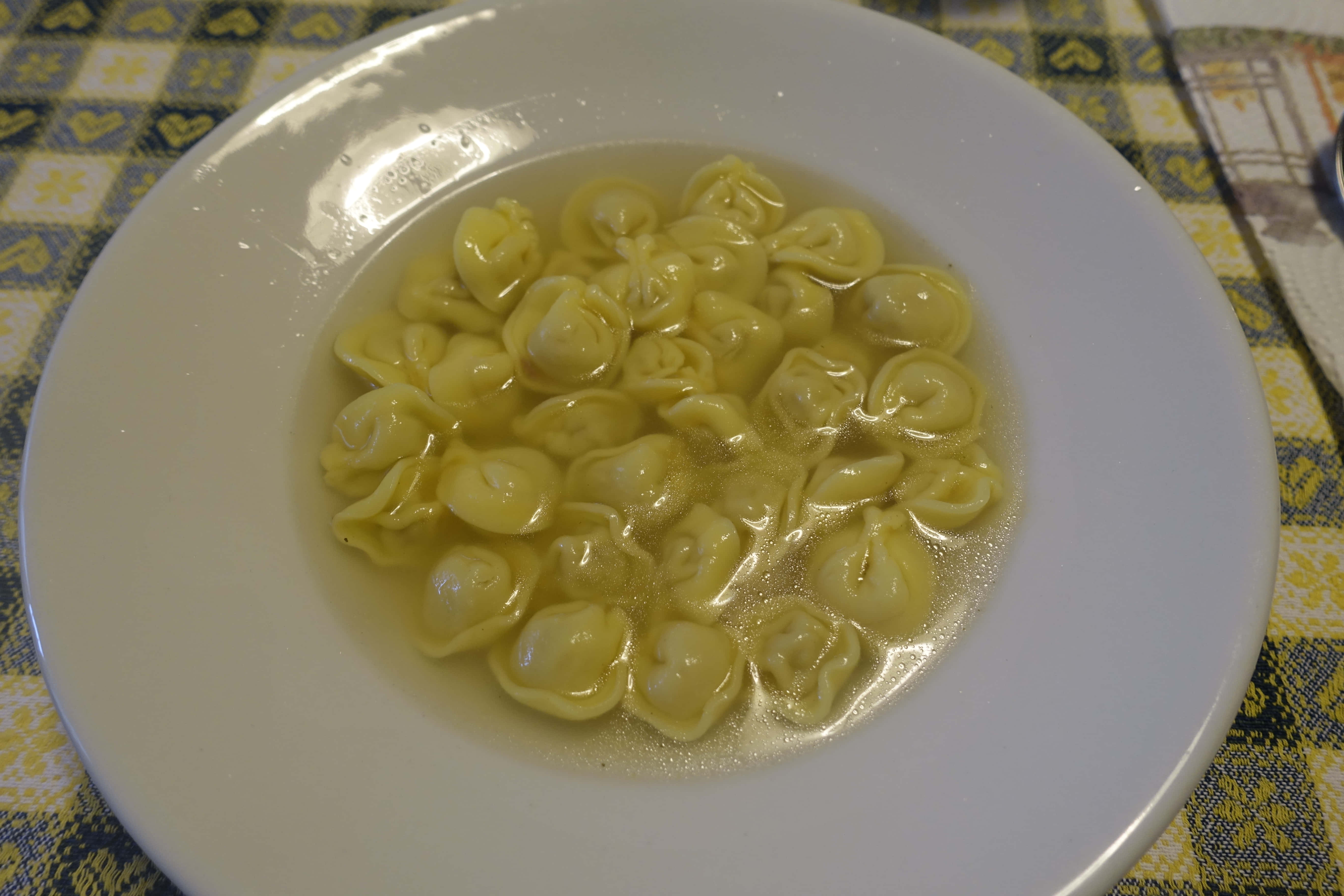 白色盤子上的義式餃子湯 壁紙 — 一個白色碗盛滿飽滿的黃色意大利餃子在清湯中，放在黃藍圖案枱布上。