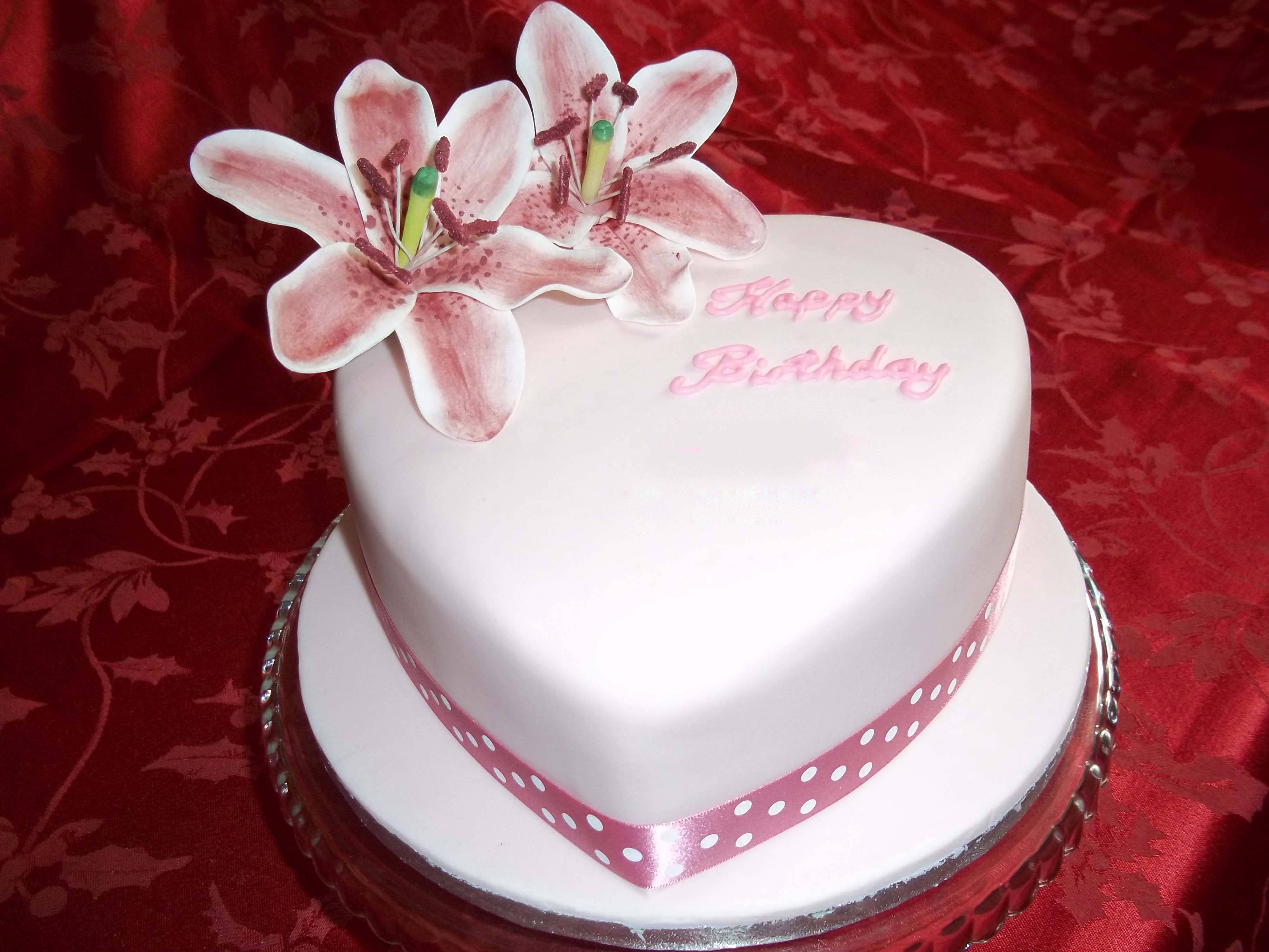 白色糖霜生日蛋糕 壁紙 — 一個心形白色生日蛋糕，飾有粉紅色百合花和波點絲帶，放在紅色圖案布料上。