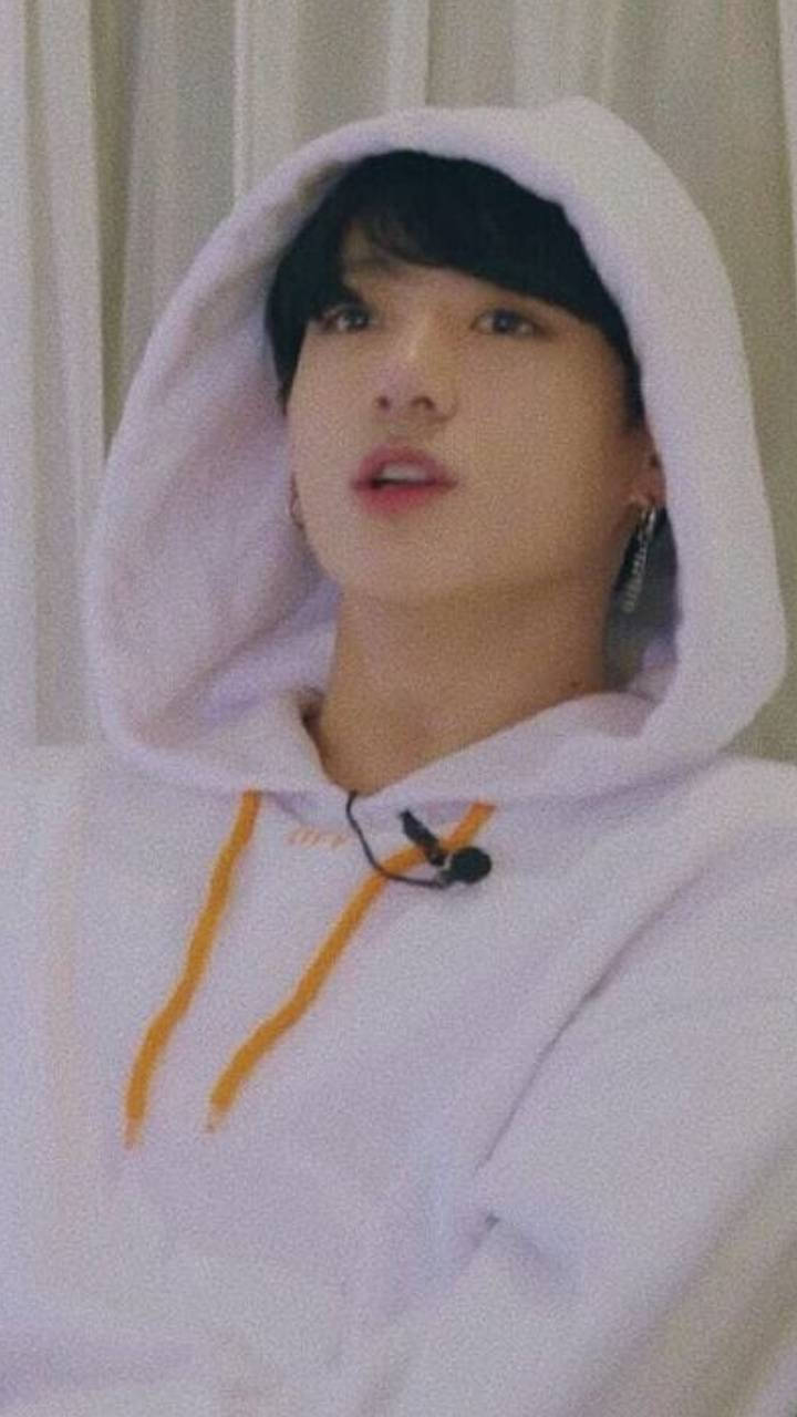 白色連帽衫 Jungkook 美學壁紙 壁紙 — 一個穿著白色連帽衫、帶橙色抽繩的年輕人，在柔軟的窗簾前抬頭仰望，表情若有所思。