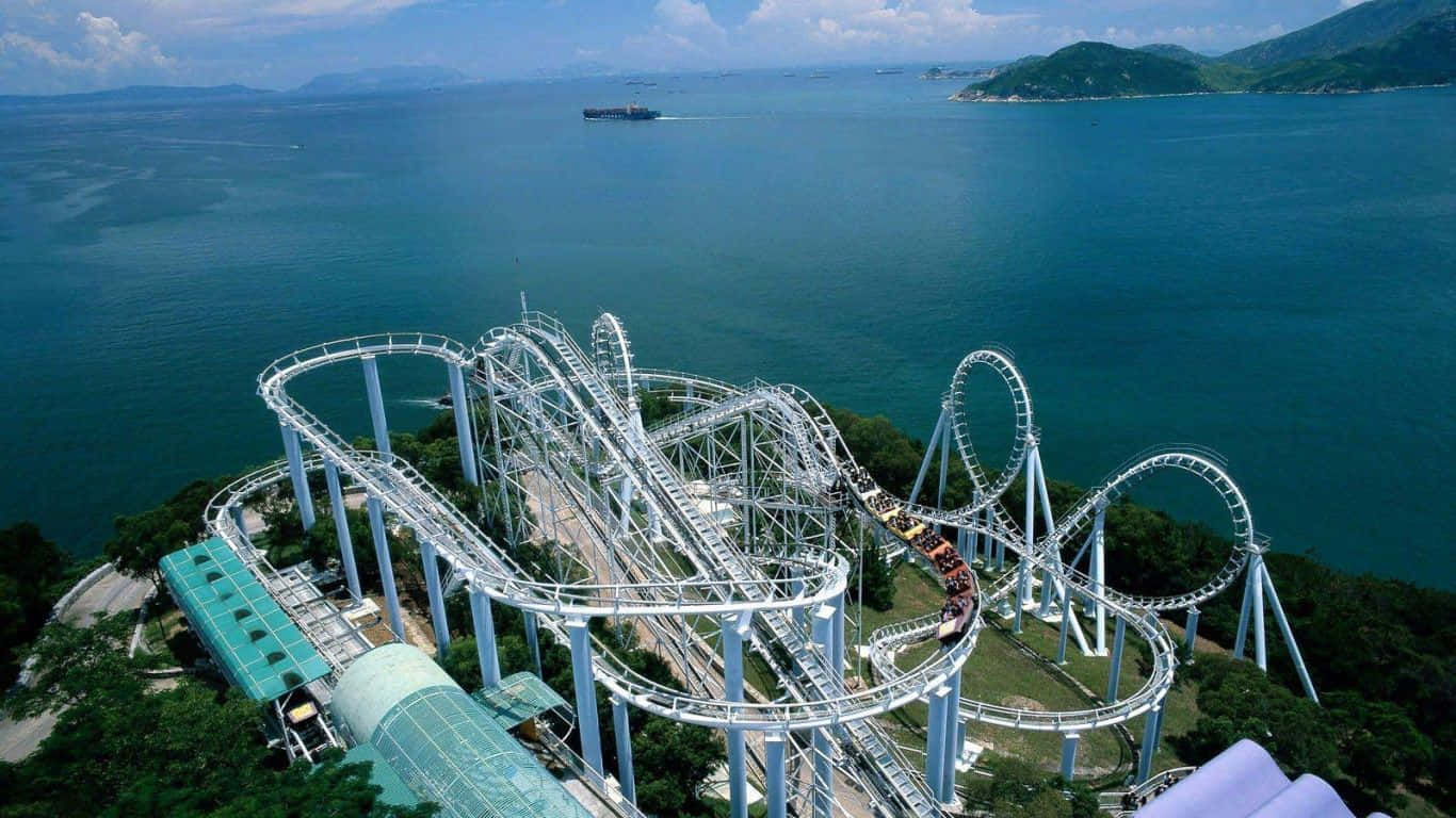 Roller Coaster 桌布 壁紙 — 白色過山車在鬱鬱蔥蔥的綠色山丘上蜿蜒曲折，旁邊是碧綠的海洋和遠處島嶼的鳥瞰圖。
