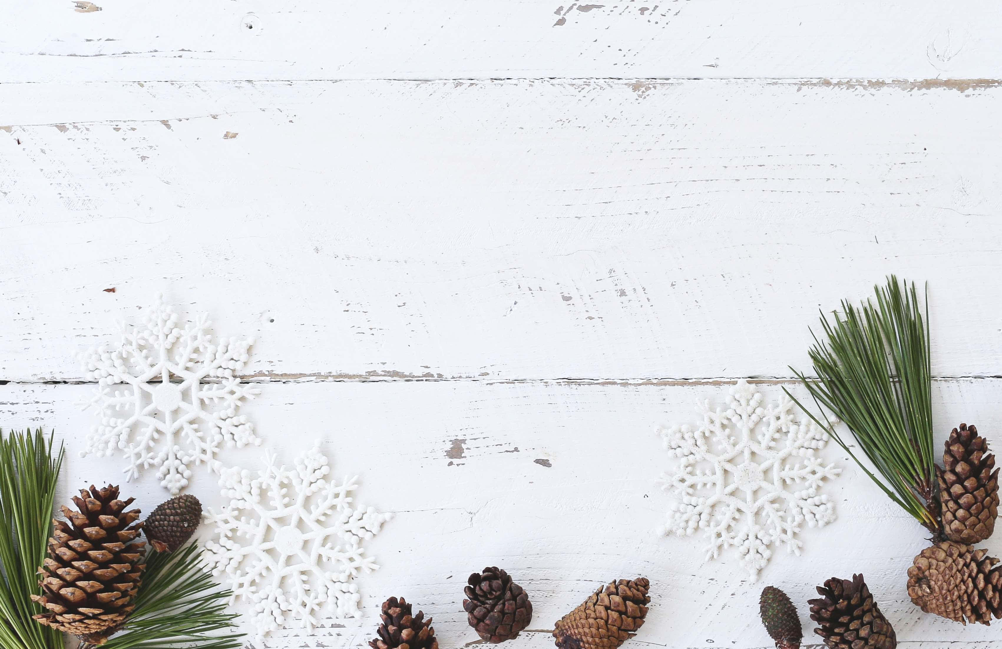 Rustic Christmas 桌布 壁紙 — 白色雪花裝飾品、松果和綠色松針排列在風化的白色木質表面上。