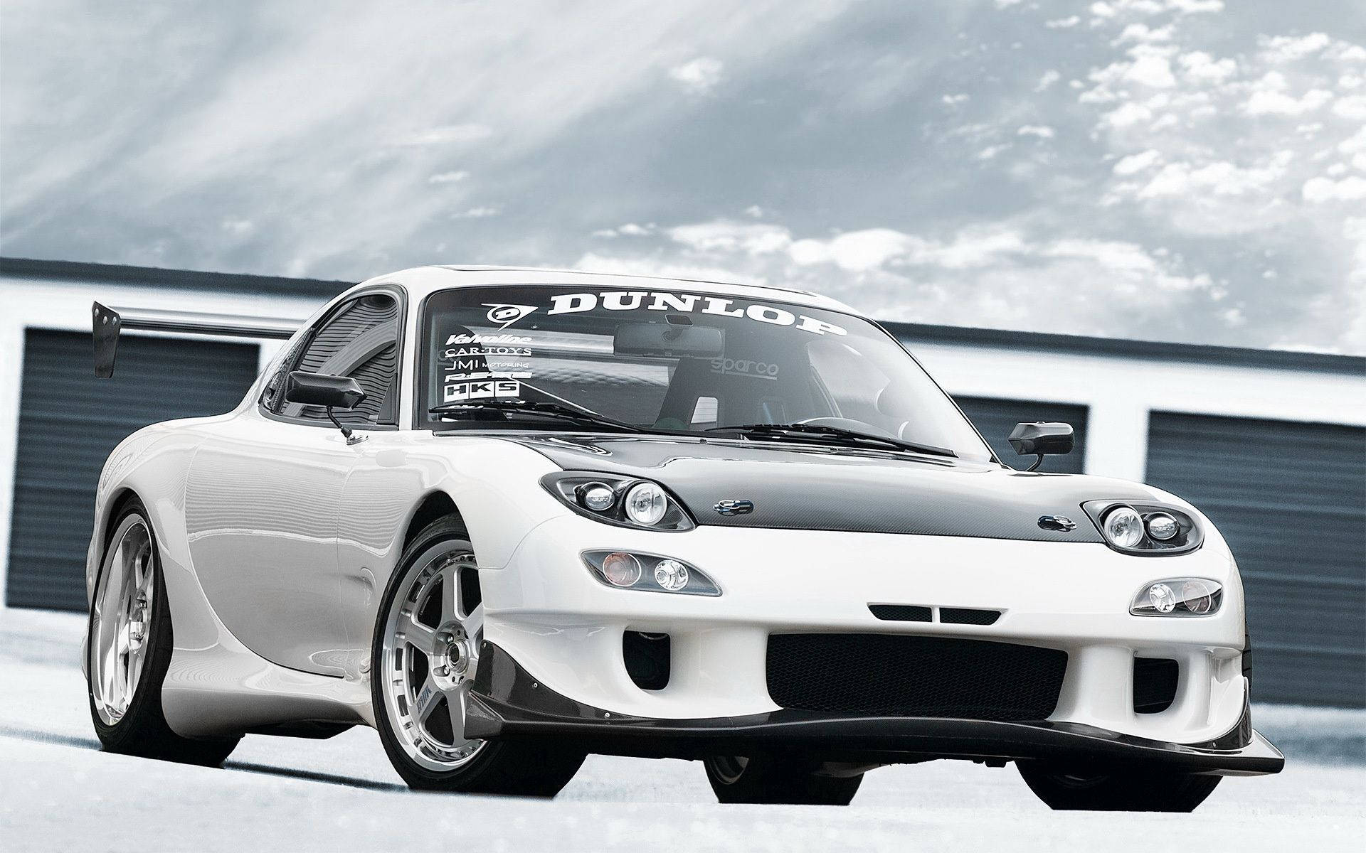 白色萬事得RX-7跑車，帶有黑色引擎蓋和賽車貼紙，停泊在車庫前。