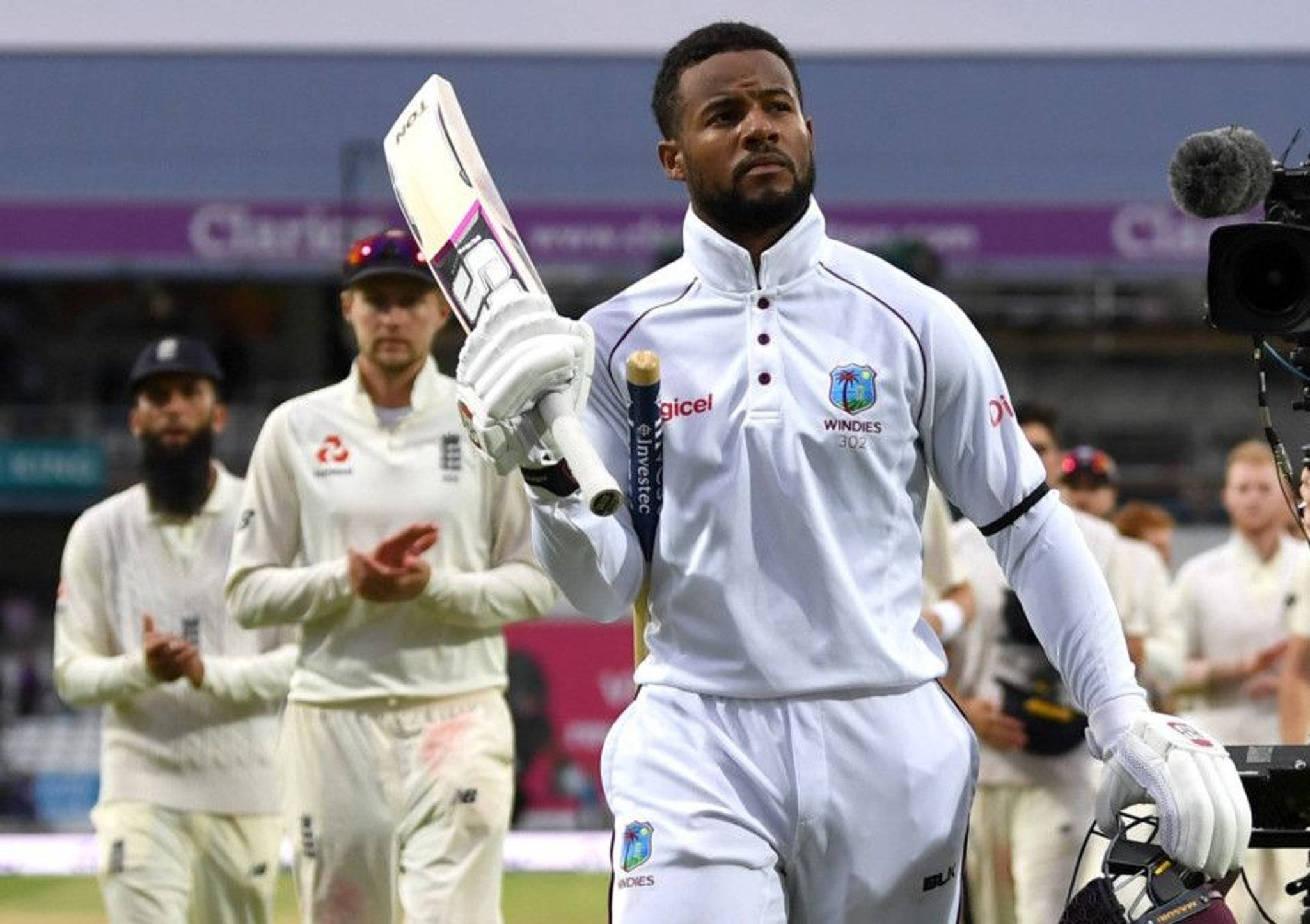 白衣中的優雅 — Shai Hope 壁紙 — 一名西印度群島板球球員身穿白色制服，高舉球棒，身後隊友在體育場上鼓掌。