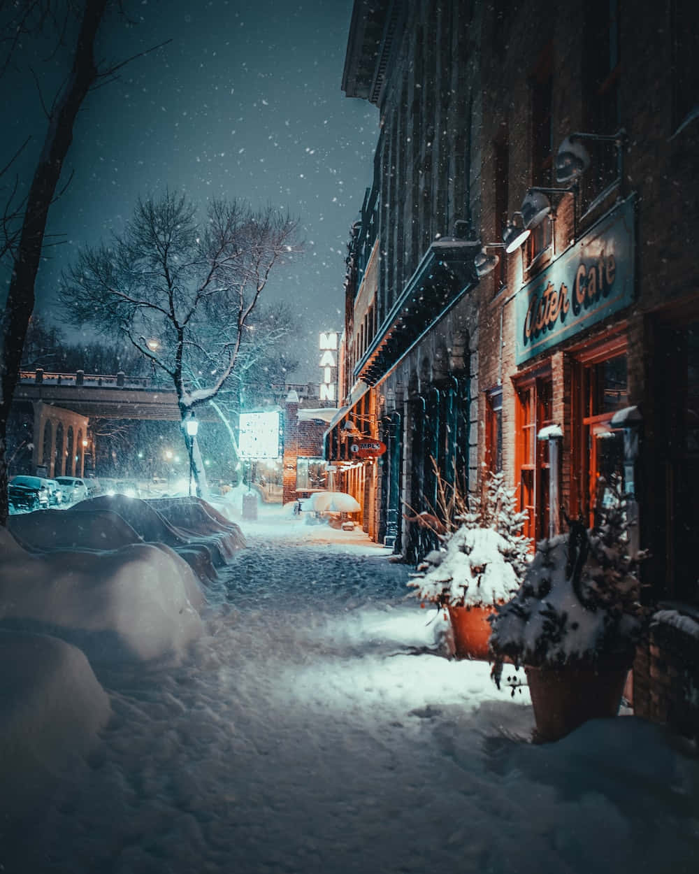 Snowy Road 桌布 壁紙 — 白雪覆蓋寧靜的城市街道，夜晚溫暖的燈光從商店櫥窗中閃爍，街燈照亮飄落的雪花。