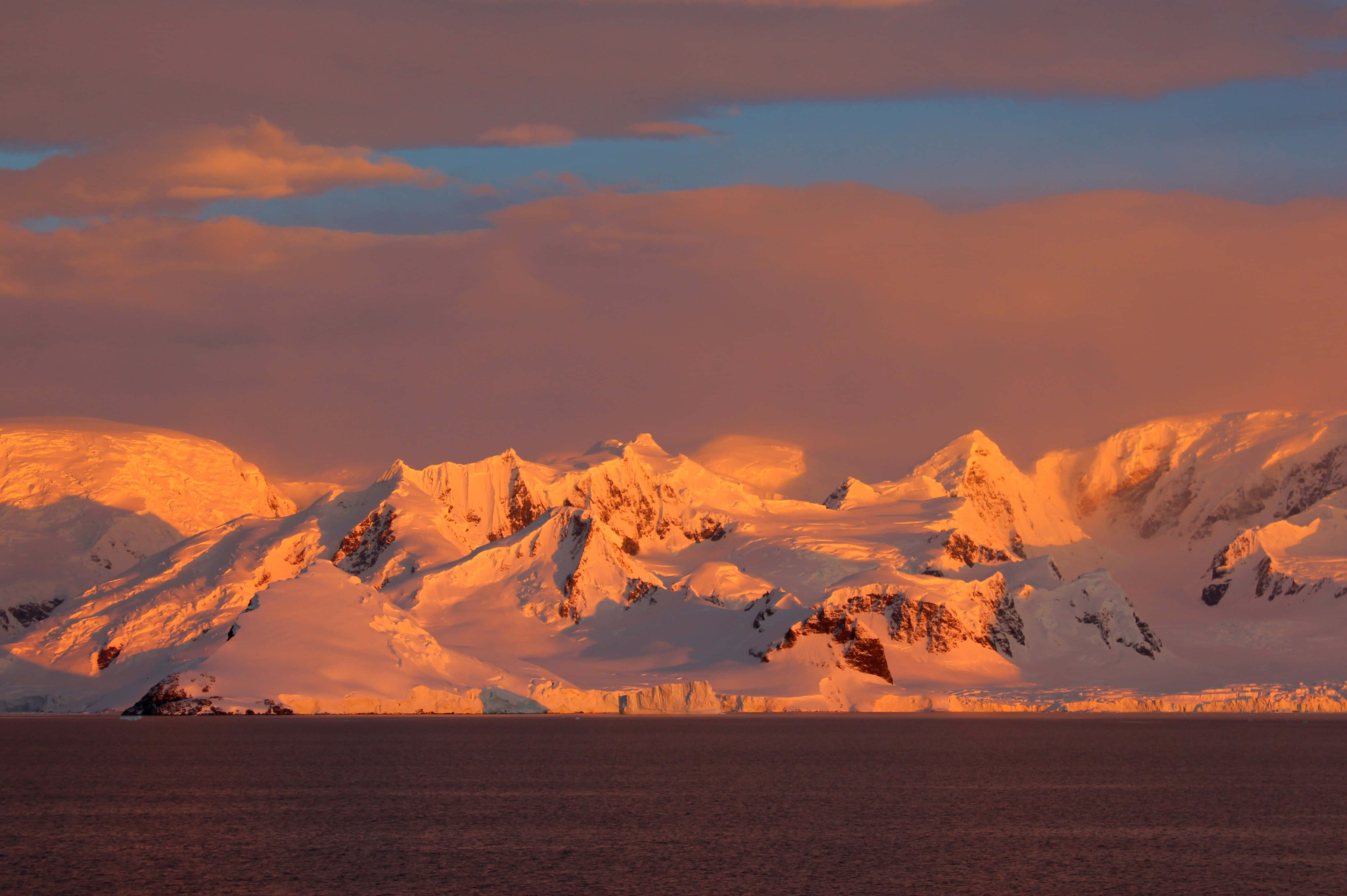 Antarctica 桌布 壁紙 — 白雪覆蓋的南極山脈在溫暖的橙色光芒中閃耀，日落時分雲朵在平靜的海面上飄移。