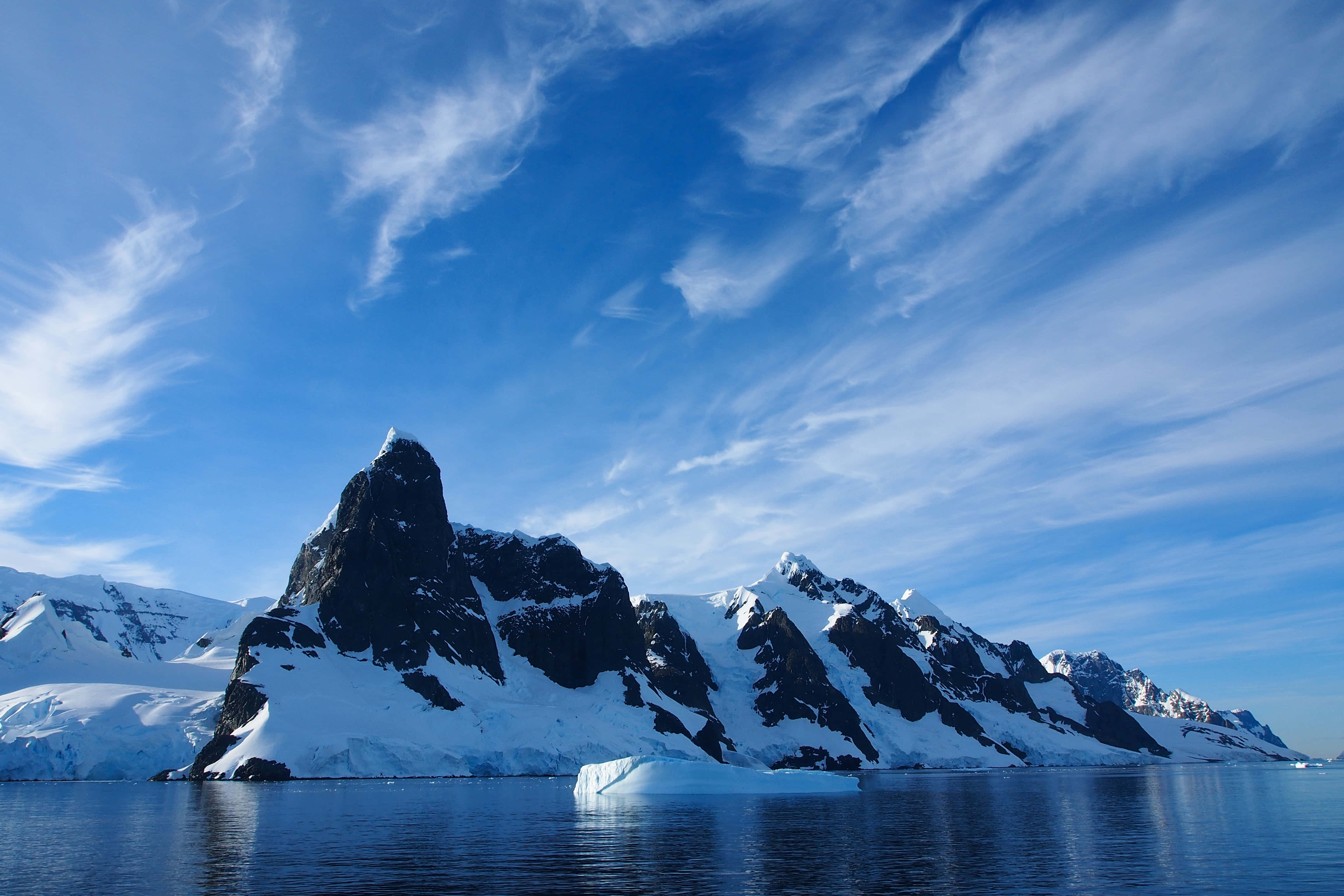 Antarctica 桌布 壁紙 — 白雪覆蓋的南極山脈從平靜的藍色海水中陡峭升起，天空色彩斑斕，點綴著縷縷薄雲。