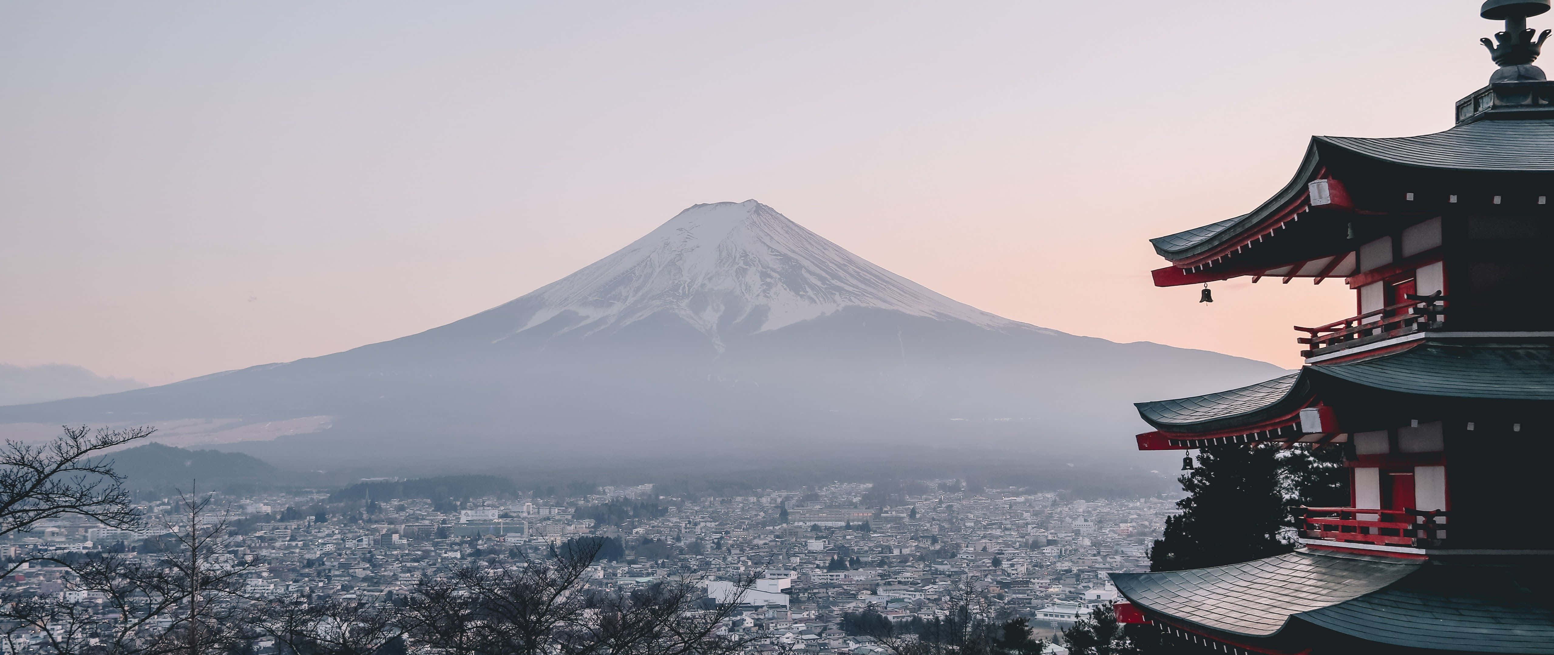 4K Pc Monitor 桌布 壁紙 — 白雪覆蓋的富士山在霧氣瀰漫的城市景觀後方雄偉地升起，一座傳統日本寶塔矗立在黎明的前景中。
