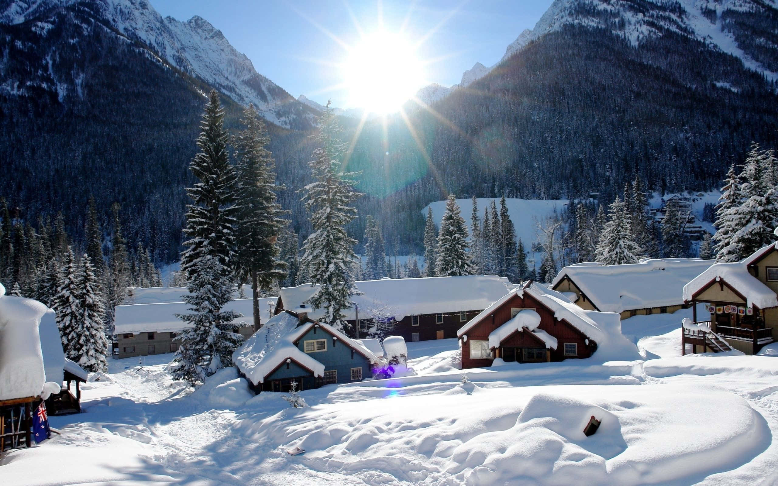 Snowy Village 桌布 壁紙 — 白雪覆蓋的小屋坐落在陽光明媚的阿爾卑斯山谷中，周圍有高聳的常綠樹木和崎嶇的山脈。