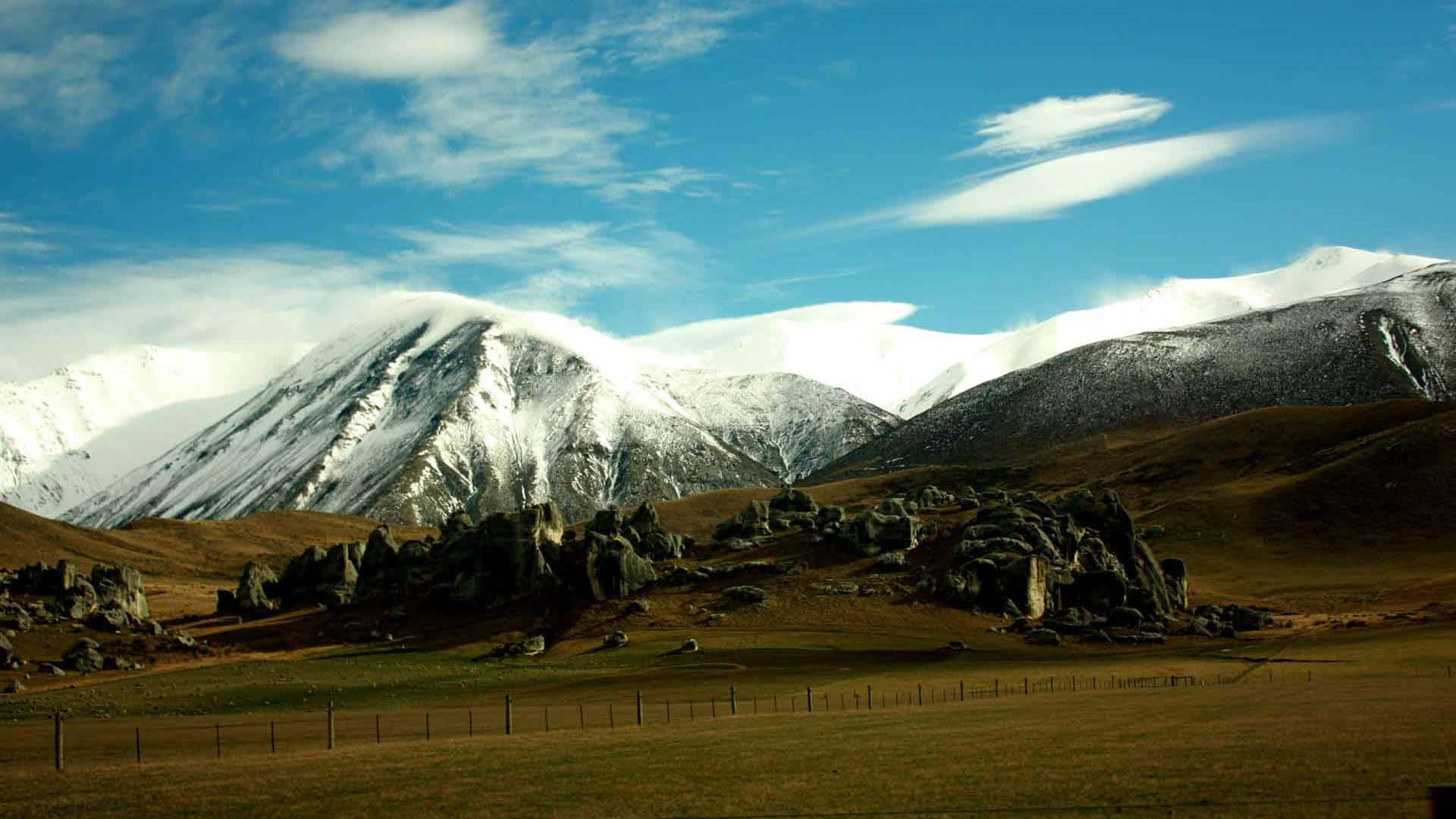 New Zealand 桌布 壁紙 — 白雪覆蓋的山峰在崎嶇的岩石地帶後方雄偉地聳立，天空湛藍，飄著縷縷輕雲。
