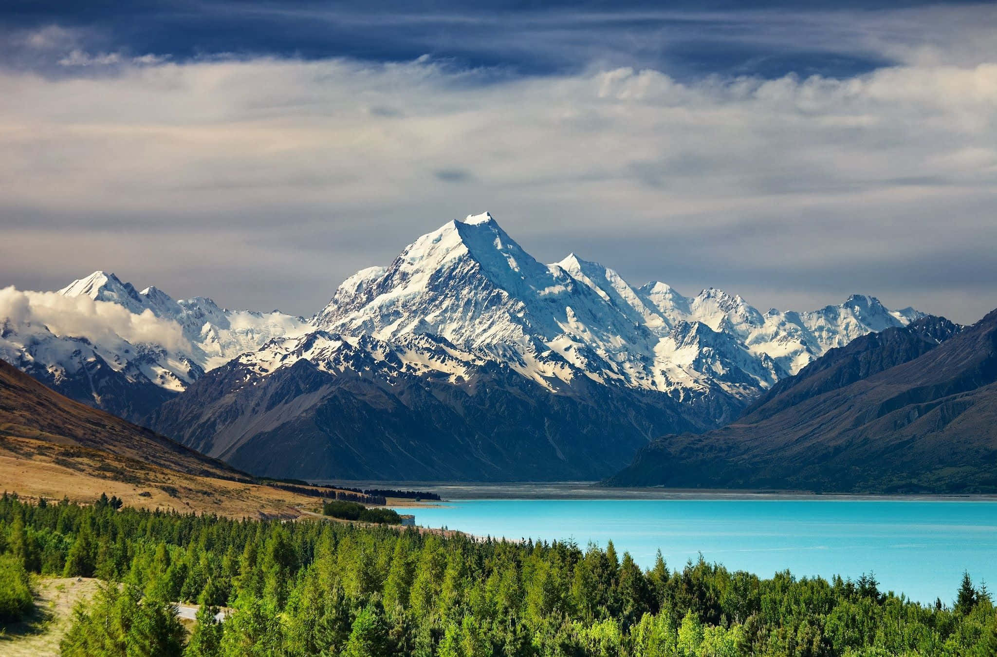 New Zealand 桌布 壁紙 — 白雪覆蓋的山峰在戲劇性的多雲天空下，雄偉地聳立在一個充滿活力的綠松石色湖泊背後，周圍環繞著茂密的綠色森林。