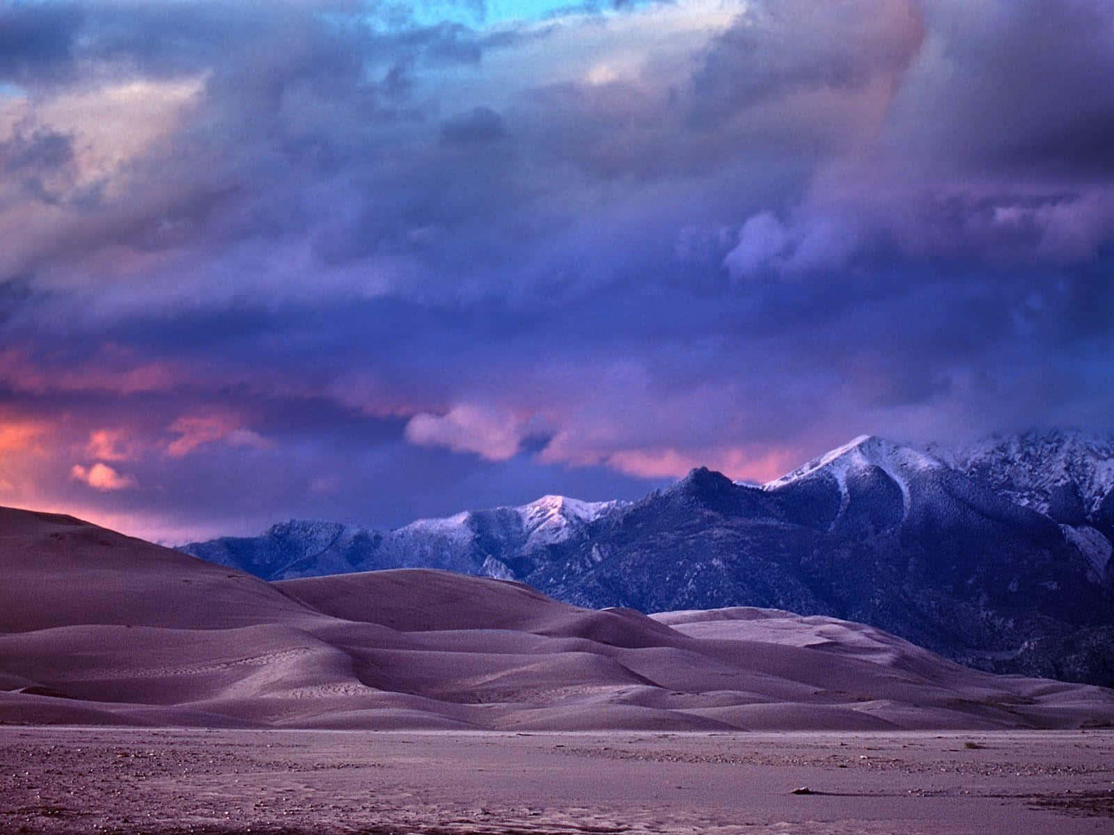 Sand Dunes 桌布 壁紙 — 白雪覆蓋的山峰在連綿起伏的沙丘後方升起，映襯著戲劇性的紫色和粉紅色日落天空。