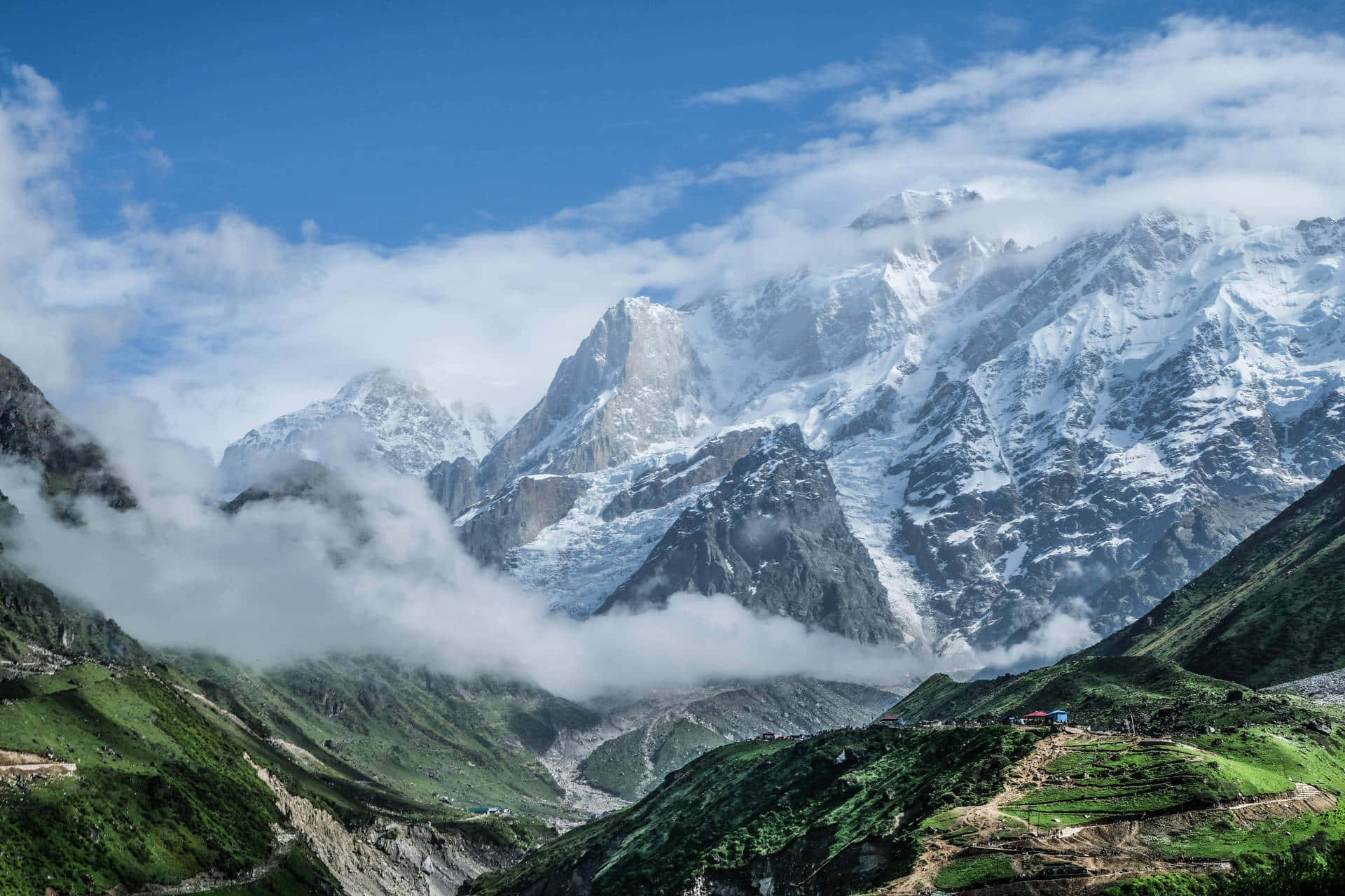 Kedarnath 桌布 壁紙 — 白雪覆蓋的山峰穿過雲層，下方是鬱鬱蔥蔥的綠色山谷，有梯田和蜿蜒的道路。
