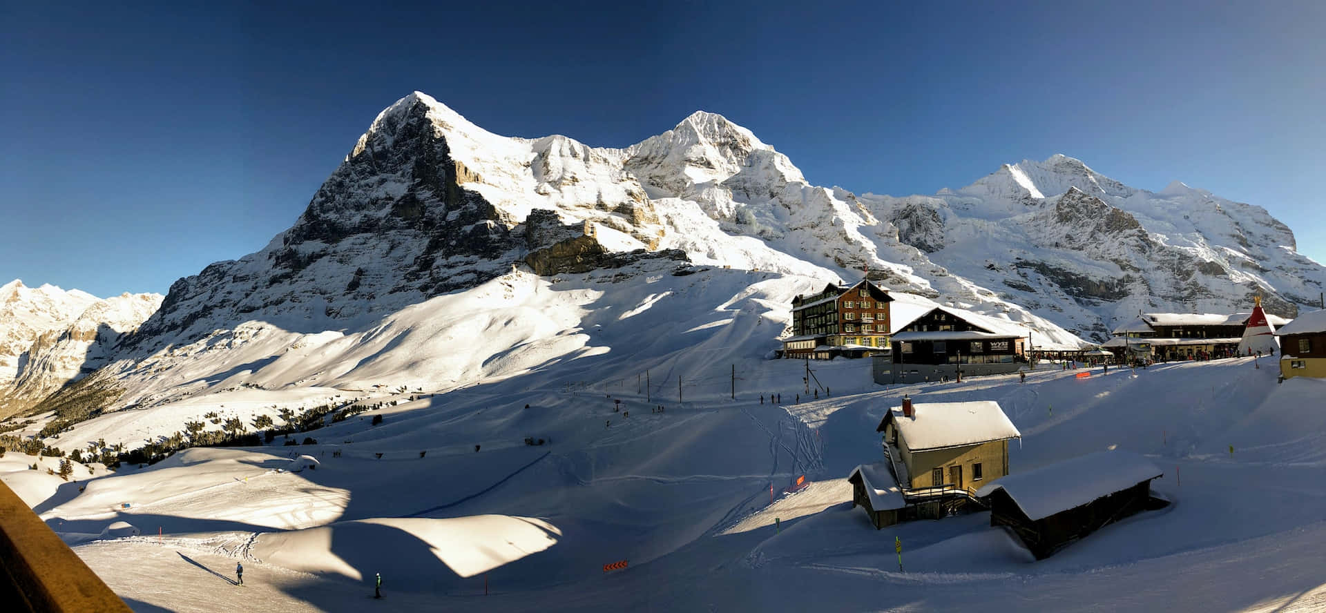 Ski Resort 桌布 壁紙 — 白雪覆蓋的山峰聳立於滑雪度假村之上，天空湛藍，滑雪者正在雪坡上滑行。