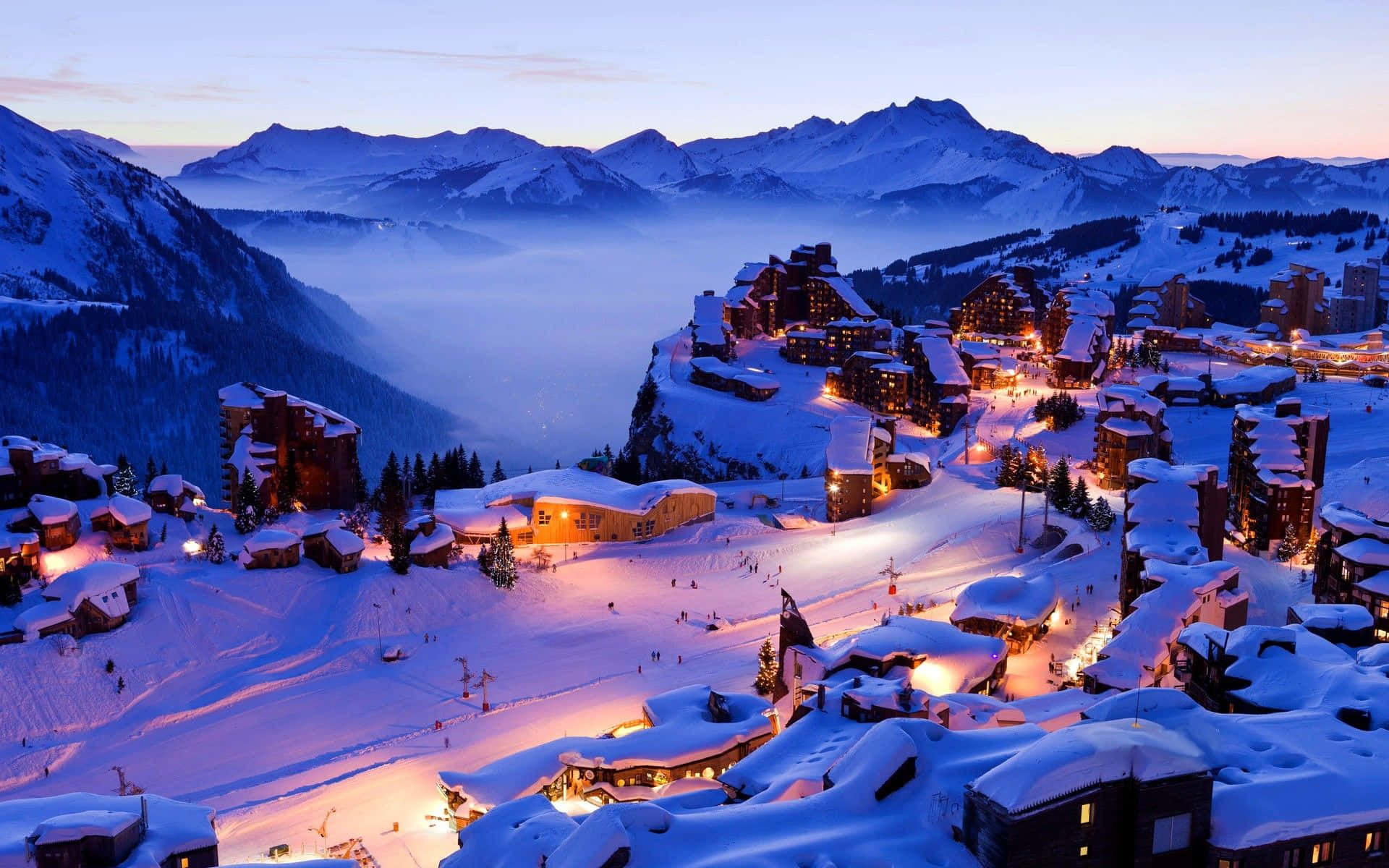 Snowy Village 桌布 壁紙 — 白雪覆蓋的山村在暮色中閃爍著溫暖的燈光，坐落在霧氣繚繞的山峰之間。