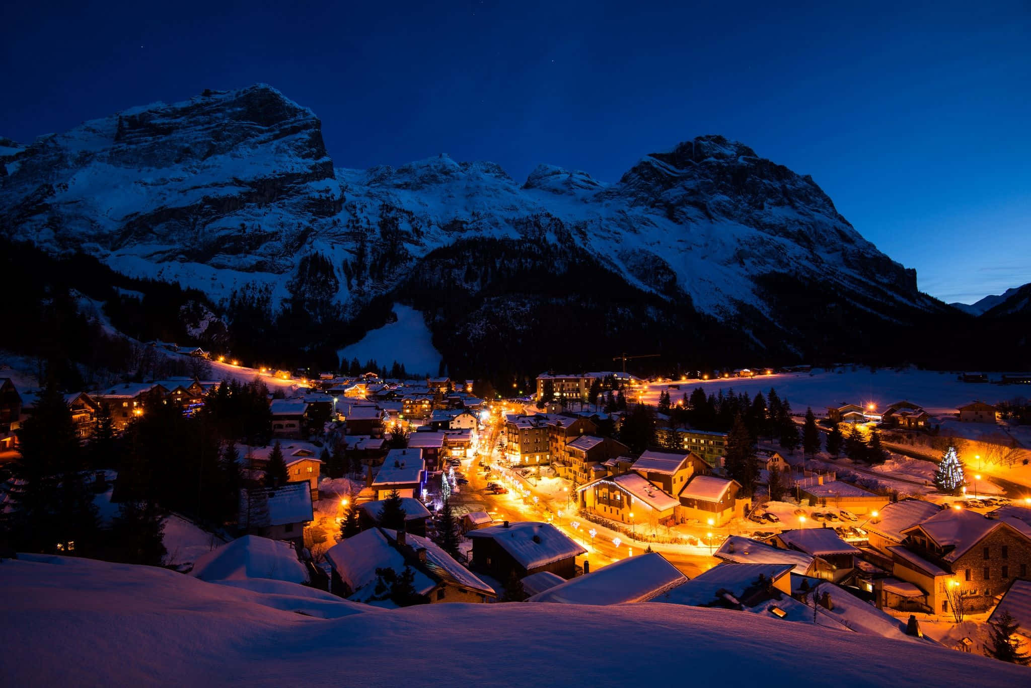 Snowy Village 桌布 壁紙 — 白雪覆蓋的山村在深藍色的暮色天空中閃爍著溫暖的燈光。