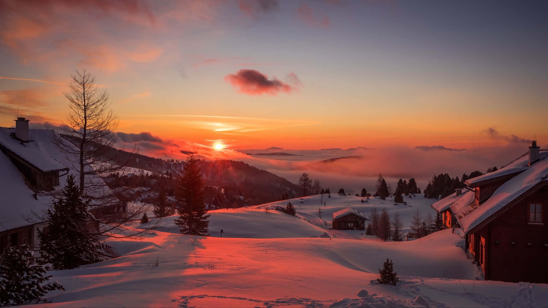 Winter Sunset 桌布 壁紙 — 白雪覆蓋的山村沐浴在熾熱的日落色調中，太陽沉沒在霧氣繚繞的山峰之下。
