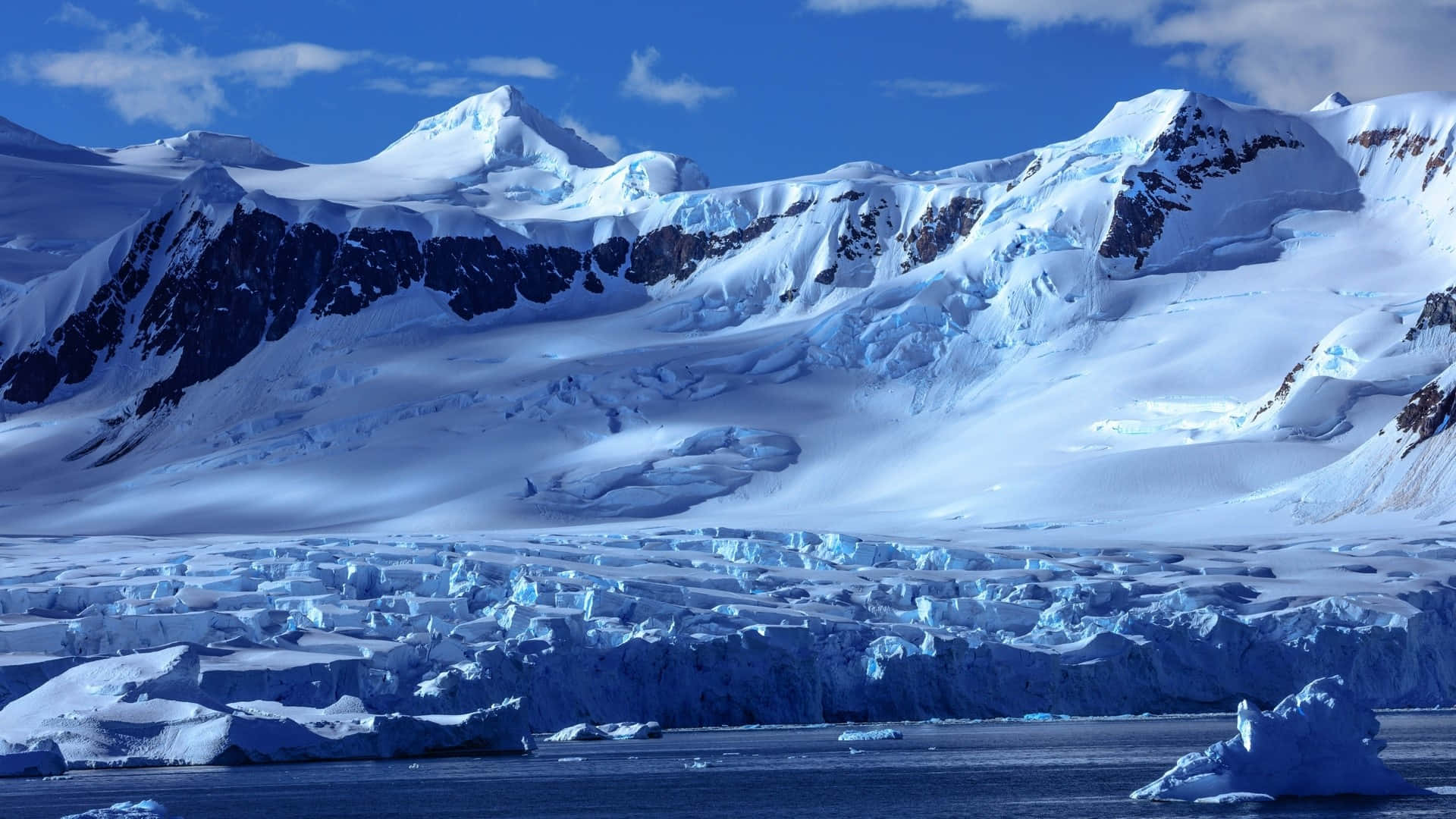 Antarctica 桌布 壁紙 — 白雪覆蓋的山脈在晴朗的藍天下聳立於點綴著參差不齊的冰山的冰凍海洋之上。