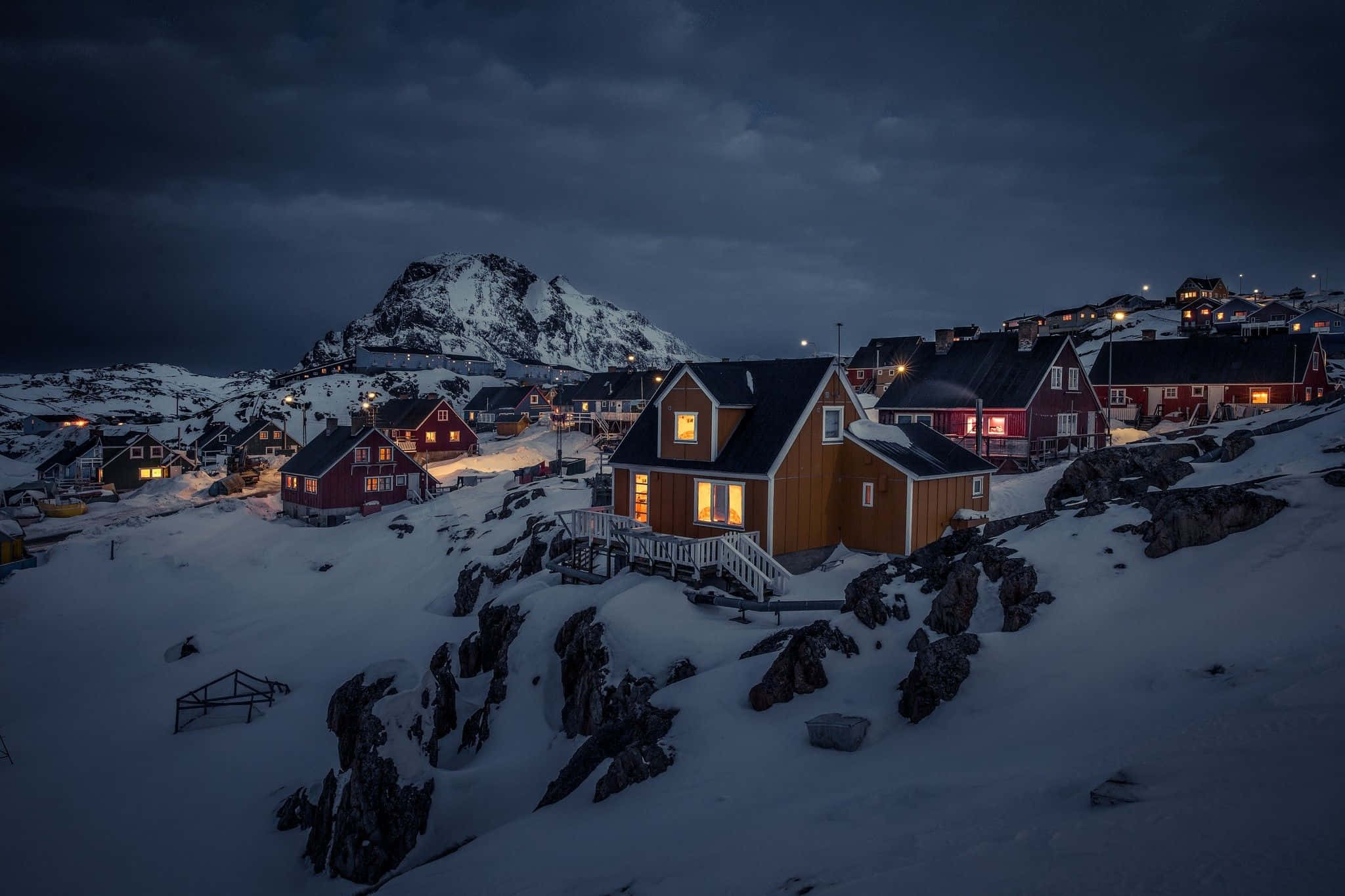 Greenland 桌布 壁紙 — 白雪覆蓋的房屋，溫暖發光的窗戶，坐落在岩石山區景觀中，在黑暗的黃昏天空下。
