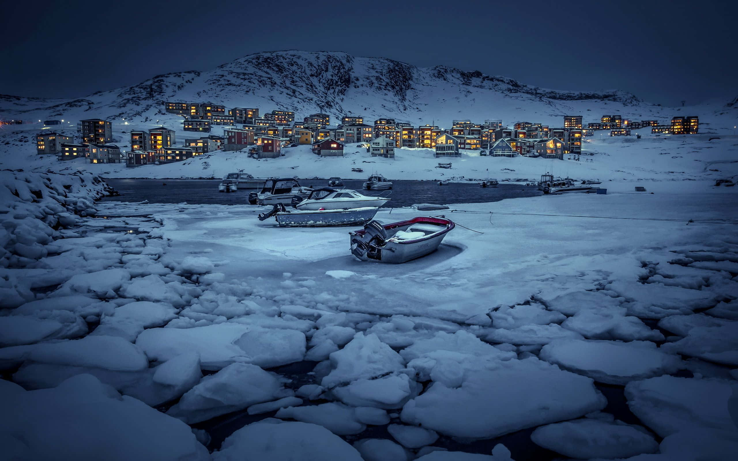 Greenland 桌布 壁紙 — 白雪覆蓋的船隻在冰凍的水面上休息，前方是一個在暮色中閃爍著溫暖燈光的山村。