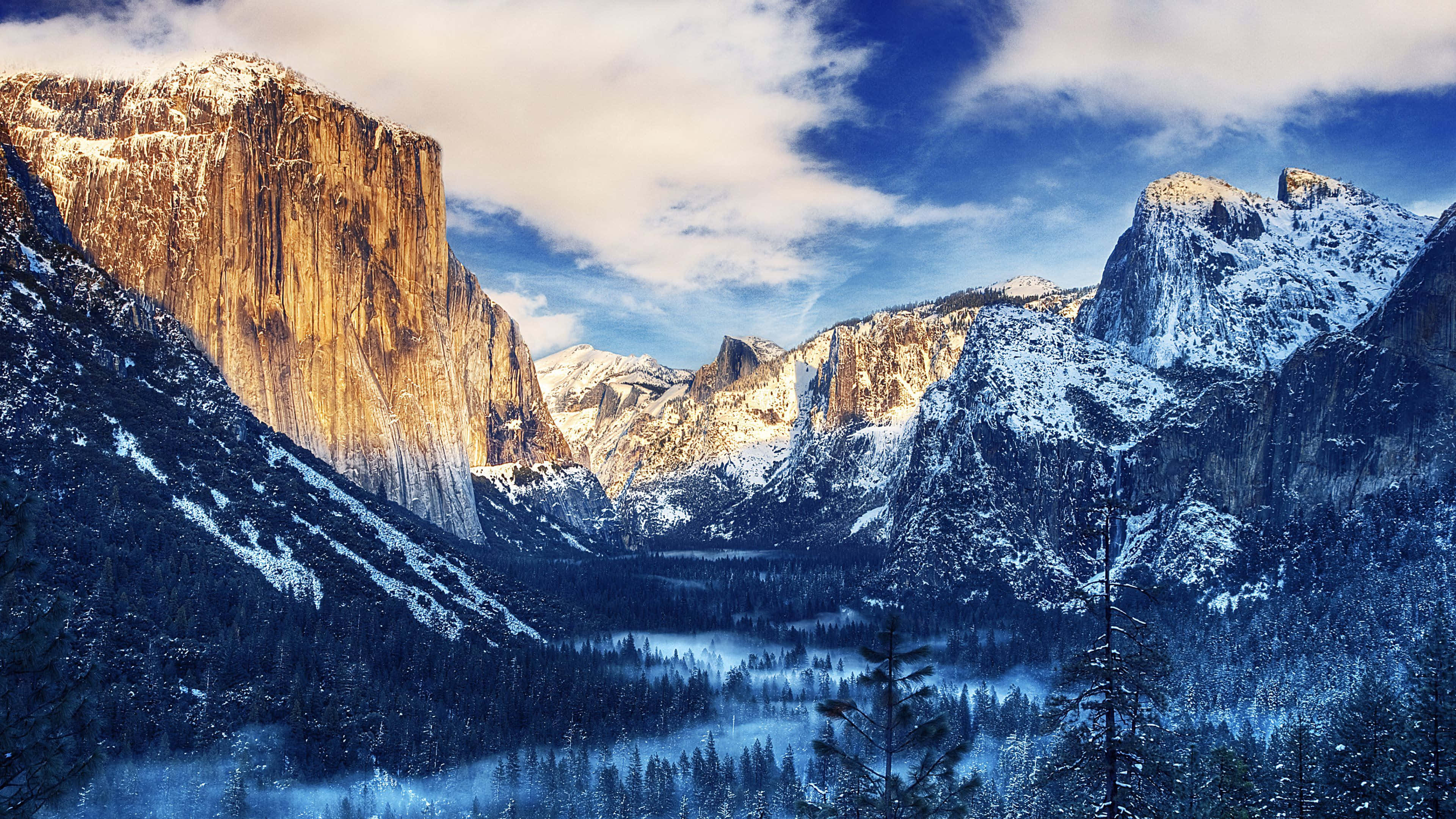 4K Display 桌布 壁紙 — 白雪覆蓋的花崗岩懸崖聳立在一個充滿常綠樹木的霧氣山谷之上，天空湛藍，白雲點綴。