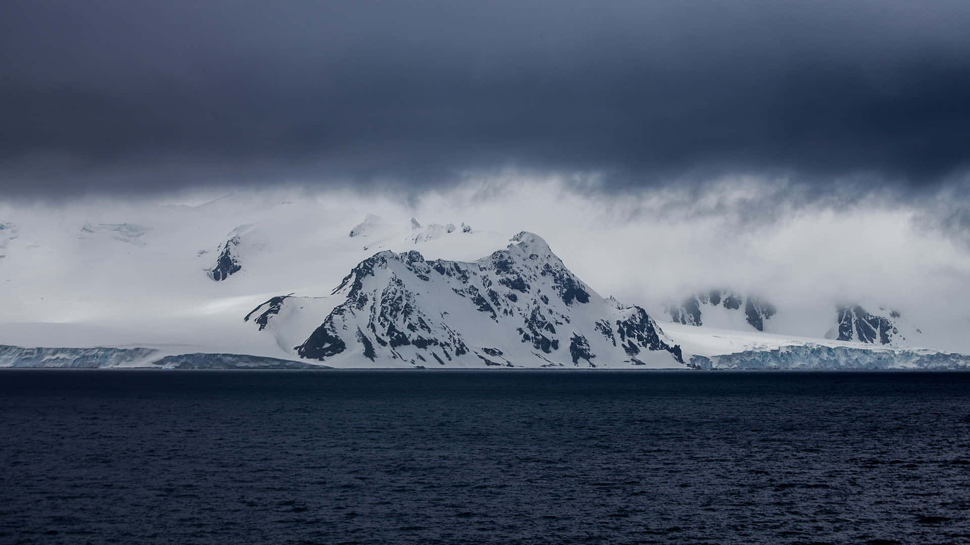 Antarctica 桌布 壁紙 — 白雪覆蓋的鋸齒狀山脈從黑暗的水域中升起，上方是低雲密佈的沉重暴風雨天空。