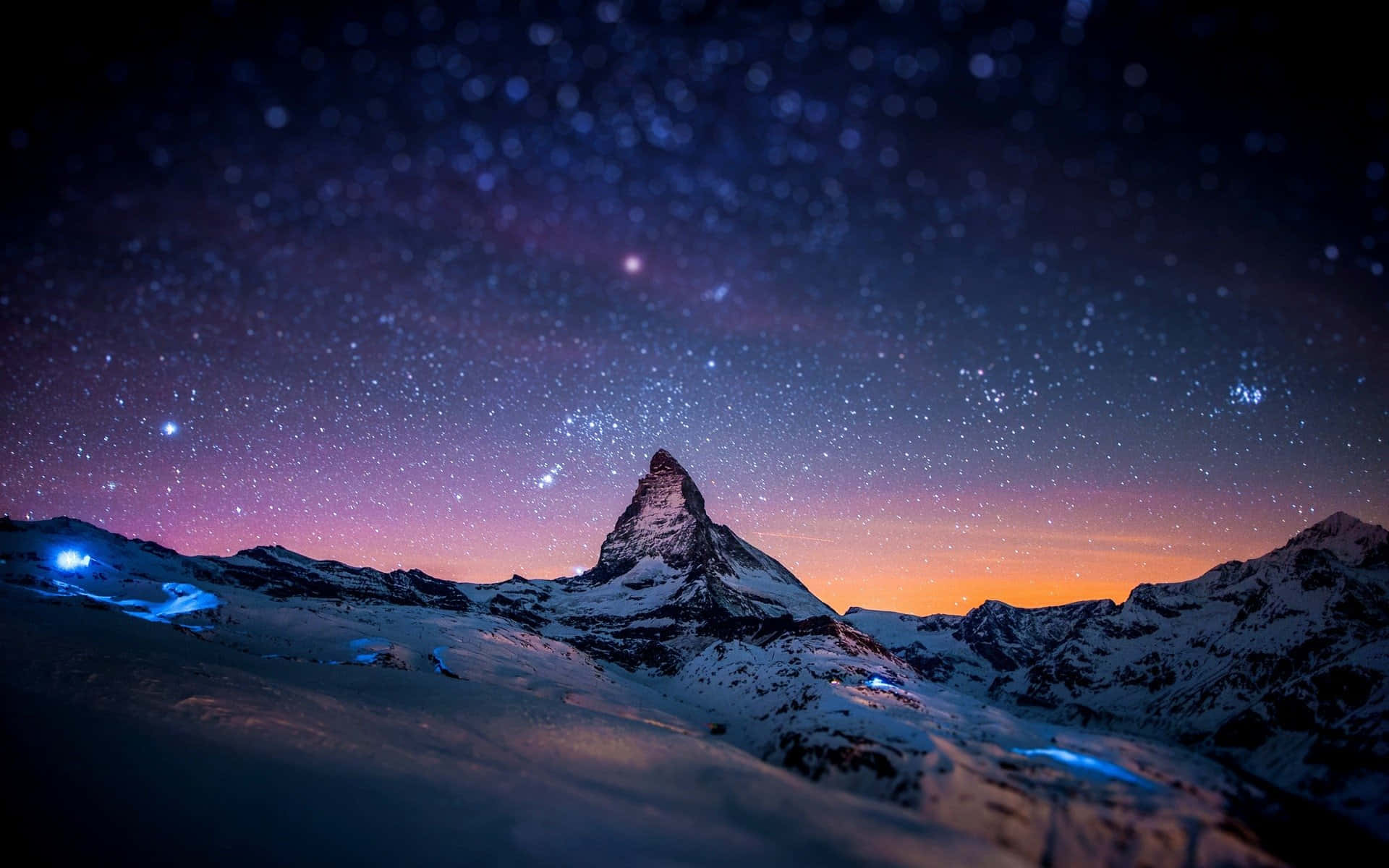 Snow Mountain 桌布 壁紙 — 白雪覆蓋的馬特洪峰在星空暮色下巍然聳立，地平線呈現紫橙色，下方有發光的藍色燈光。