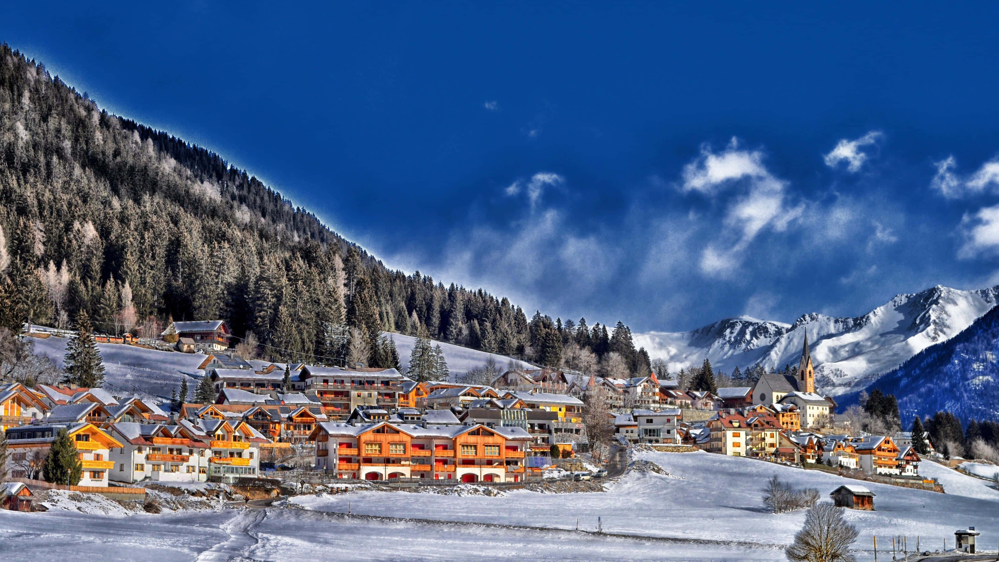 Snowy Village 桌布 壁紙 — 白雪覆蓋著一個充滿活力的阿爾卑斯山村，坐落在高聳的松樹林中，天空湛藍，雲朵點綴。