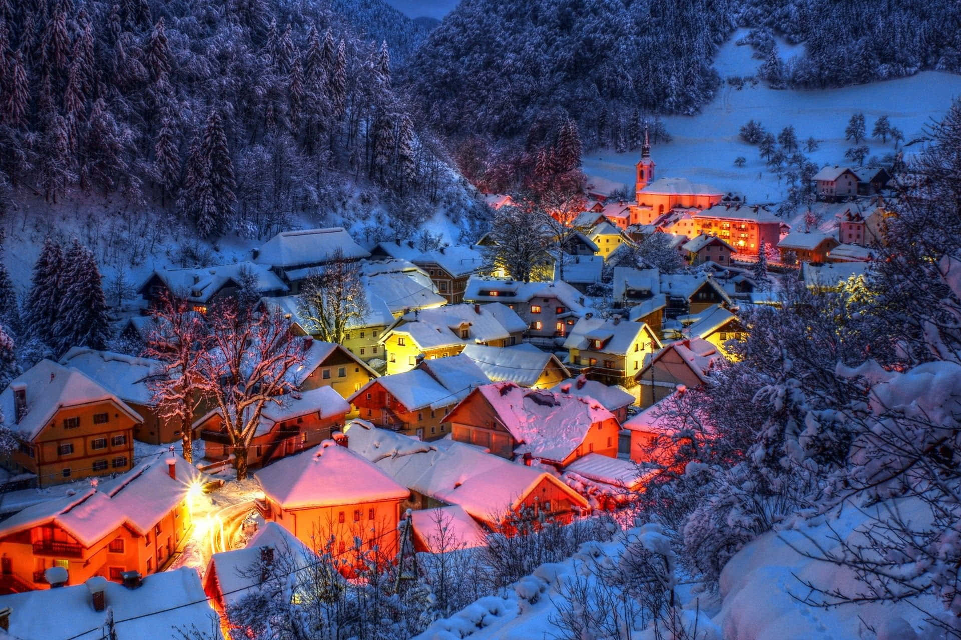 Snowy Village 桌布 壁紙 — 白雪覆蓋著一個舒適的山村，在暮色中，溫暖的金色燈光從窗戶透出，映襯著深藍色的天空。