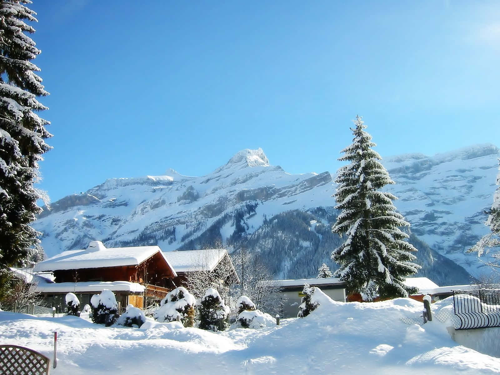 Winter Holiday 桌布 壁紙 — 白雪覆蓋著一個舒適的阿爾卑斯山村，坐落在晴朗藍天下高聳、陽光明媚的山腳下。