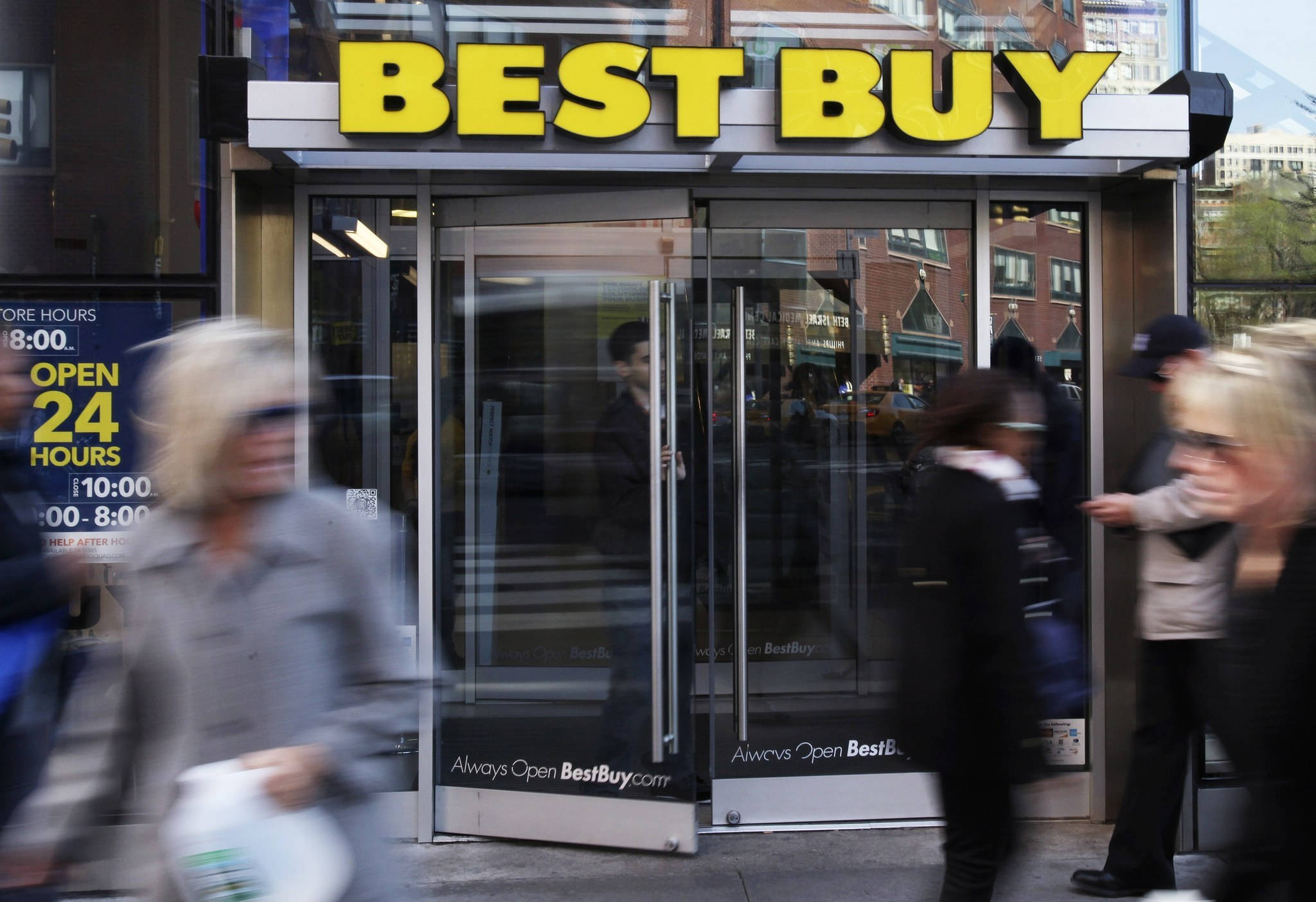 百思 buy 過往行人 壁紙 — 模糊的行人匆匆經過Best Buy商店入口，入口有醒目的黃色標誌和玻璃門，反射出城市建築。
