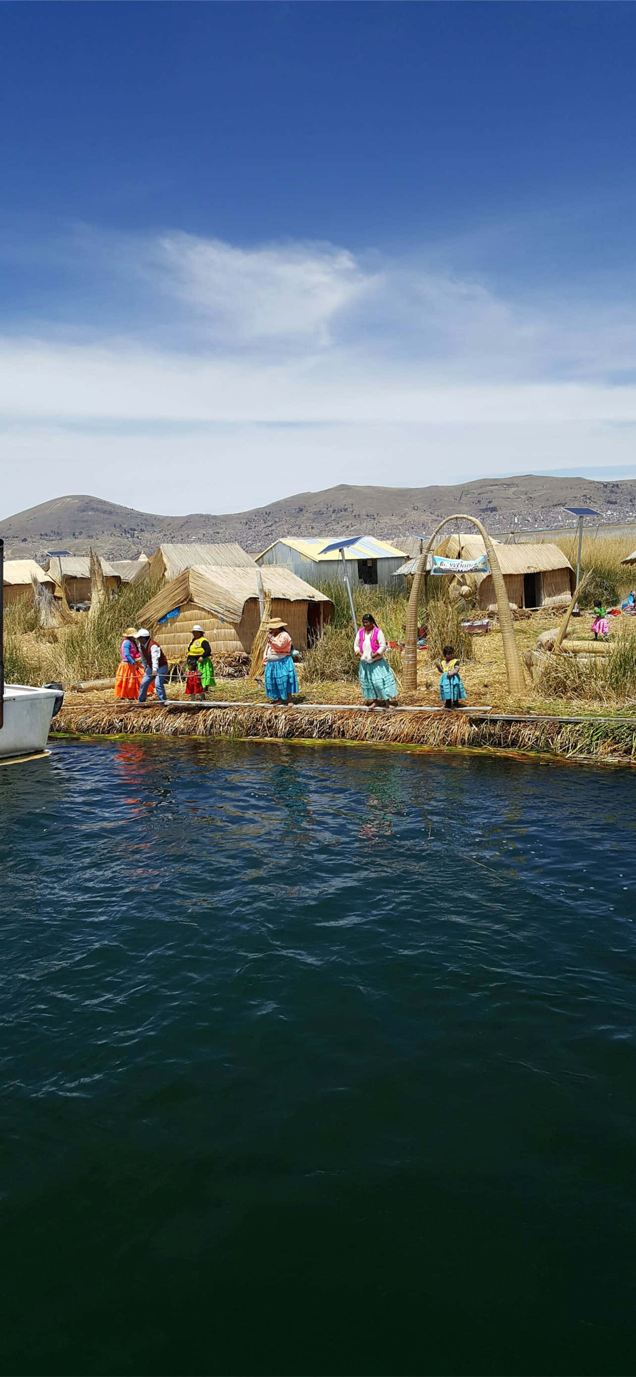 的喀喀湖上的烏羅浮島 壁紙 — 鮮豔的傳統房屋建在漂浮的蘆葦島上，人們穿著色彩繽紛的服裝，靠近平靜的深色水域，上方是明亮的藍天。