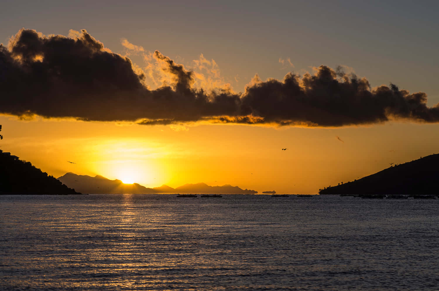 的的喀喀湖日落 壁紙 — 金色夕陽在遠山後方閃耀，將溫暖的光芒投射在平靜的水面上，上方有深色雲朵剪影。