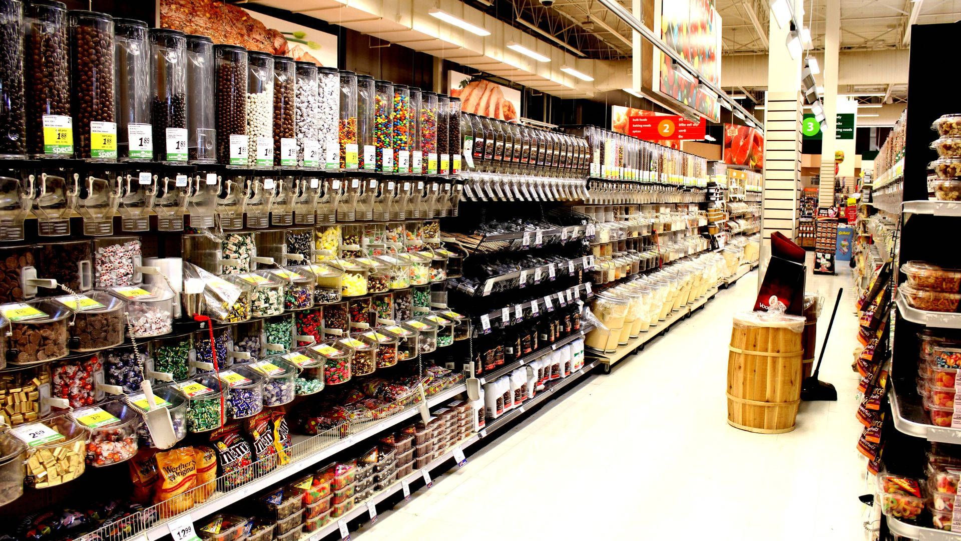 皇家印度雜貨店 壁紙 — 燈光明亮的超市通道，裝滿了透明容器和貨架上的彩色散裝糖果和小吃。