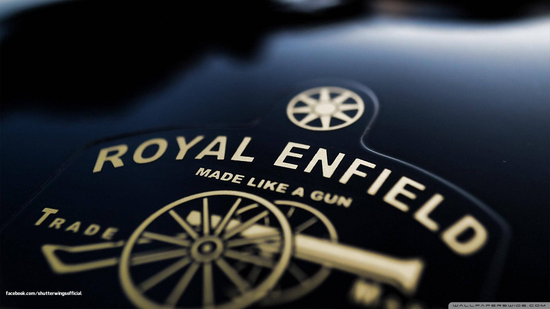 皇家恩菲德高畫質標誌與口號 壁紙 — Royal Enfield 電單車標誌的特寫，金色字體和大炮徽章在光澤黑色表面上。