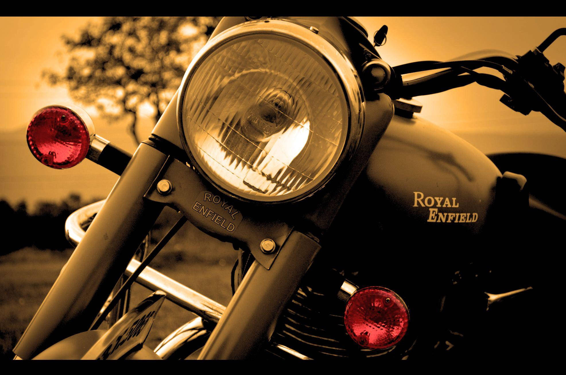 皇家恩菲爾德 HD 塞普亞調 壁紙 — 復古Royal Enfield電單車頭燈在金色日落背景下溫暖發光的特寫。