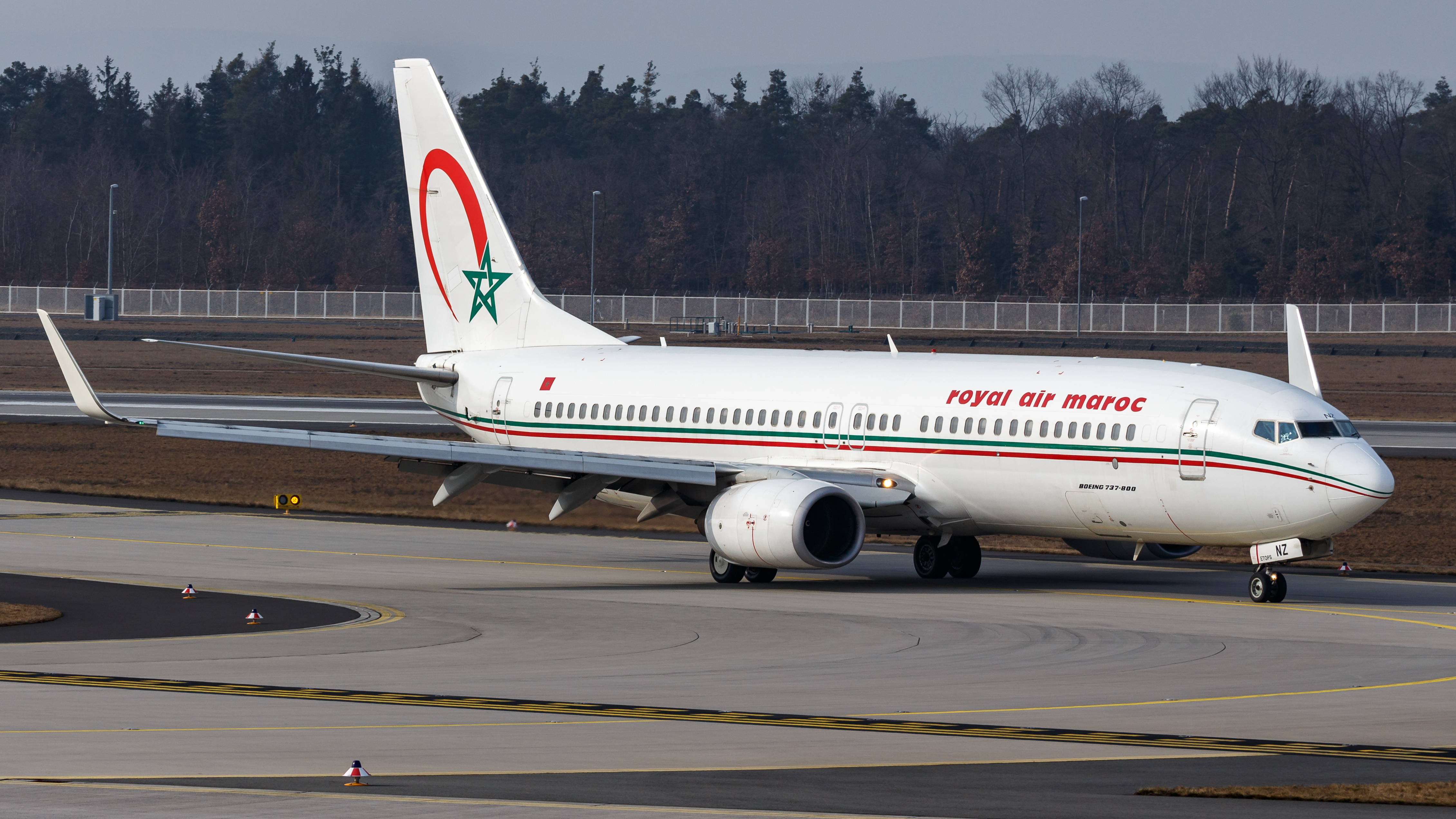 皇家摩洛哥航空與高樹 壁紙 — 一架摩洛哥皇家航空的波音737在灰色跑道上滑行，背景有光禿禿的樹木和圍欄，天空多雲。