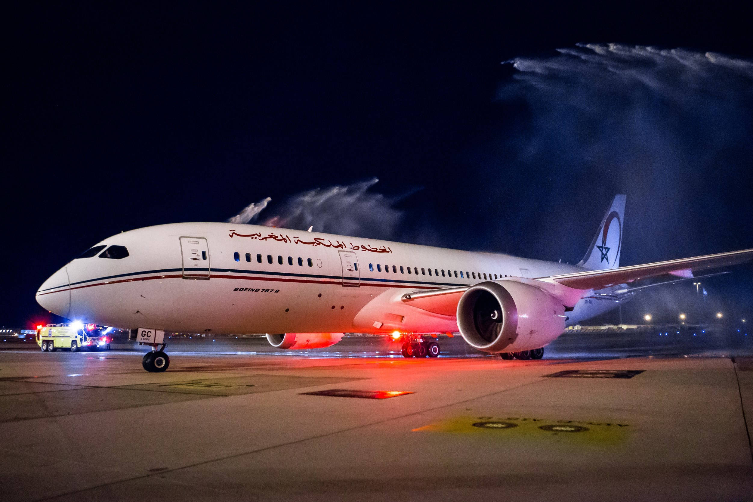 皇家摩洛哥航空飛機伴水花飛行 壁紙 — 一架白色的阿聯酋航空空中巴士A350沐浴在紅光中，水炮在夜間濕滑的停機坪上從後方噴射。